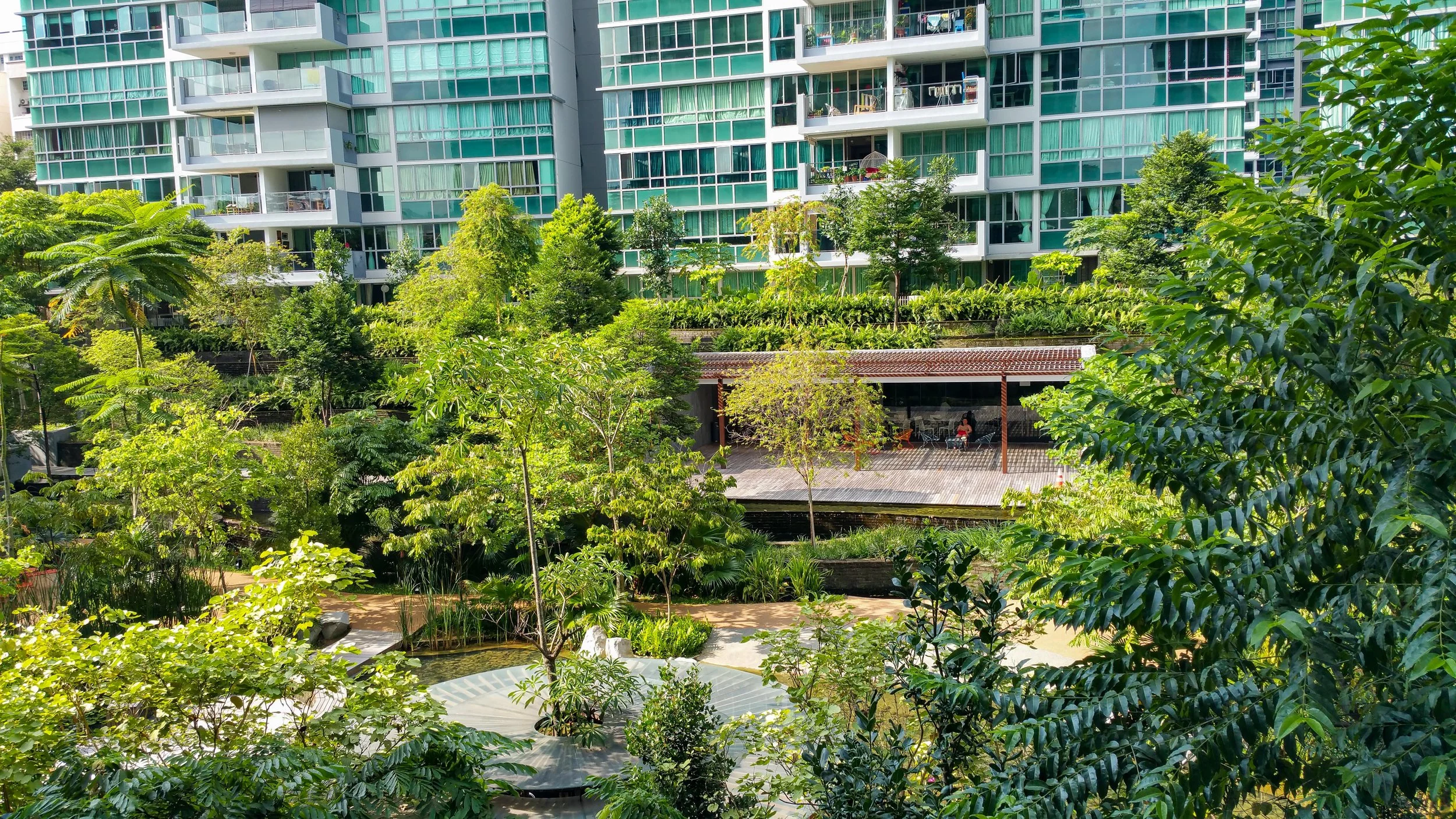 View of a lush green courtyard with various trees and plants, with modern glass apartment buildings in the background and a shaded outdoor seating area with people.