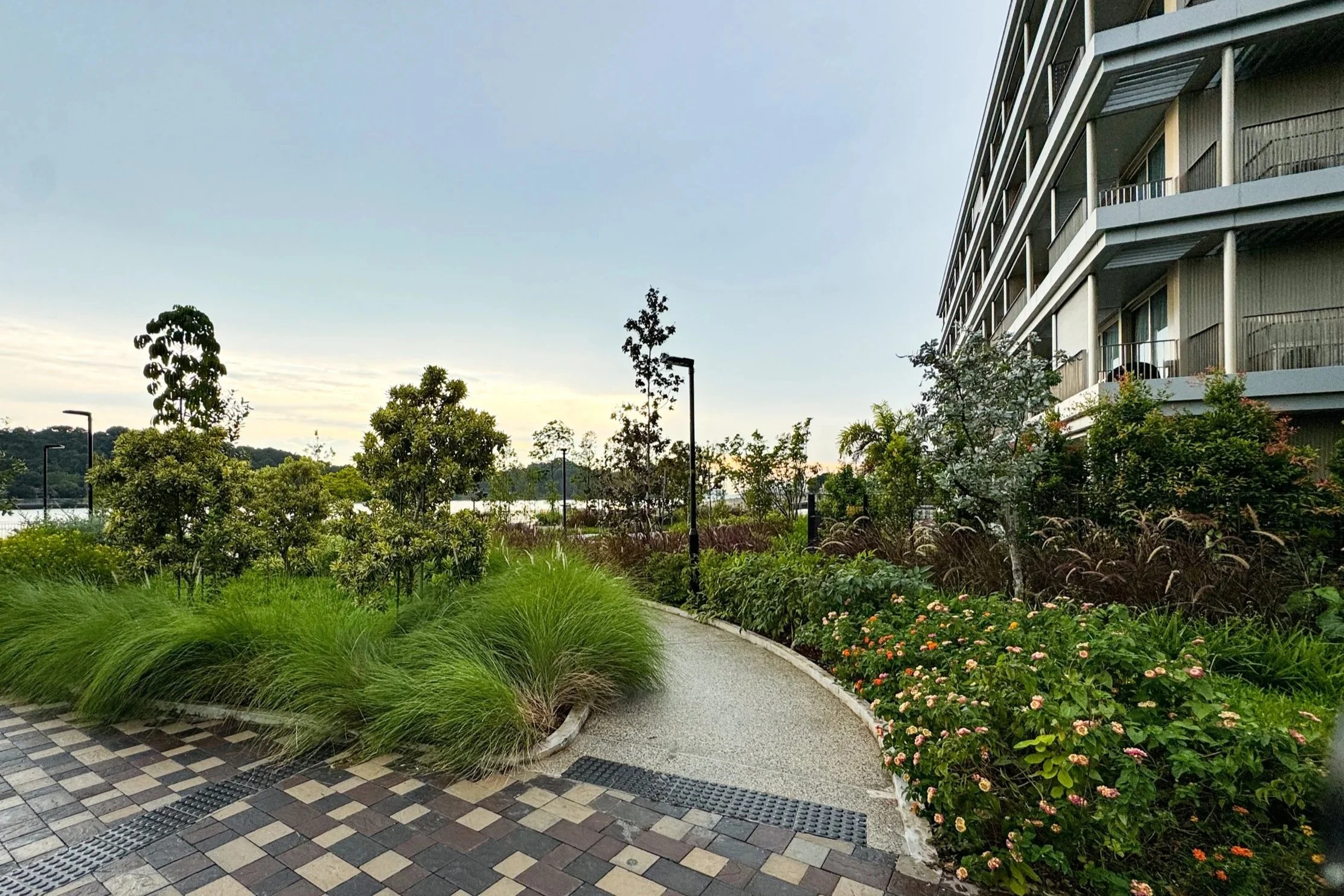 Pathway lined with green shrubs and flowering plants next to a modern multi-story apartment building with balconies, under a cloudy sky.