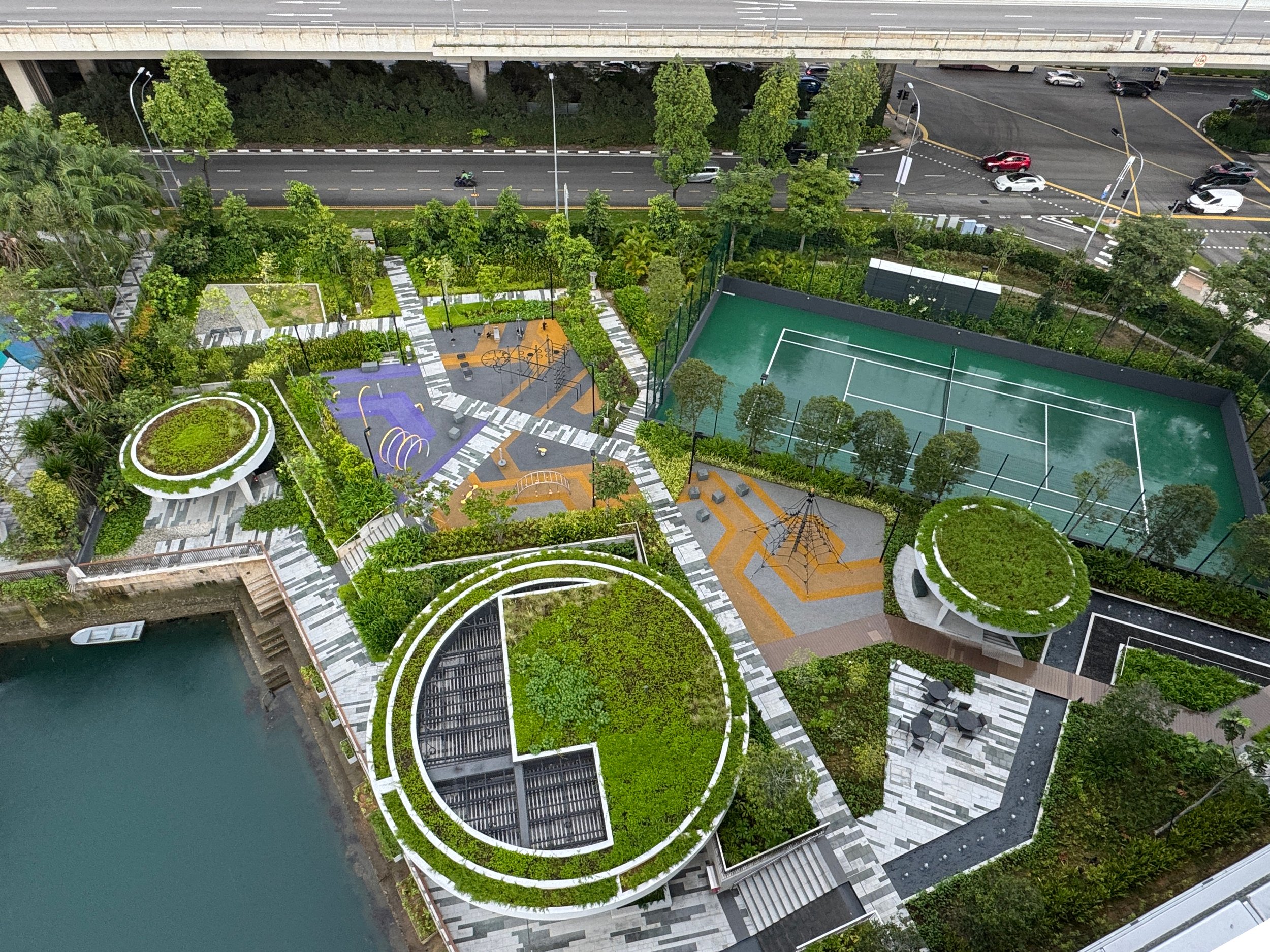 Bird's eye view of a green rooftop garden with walking paths, seating areas, and a children’s playground, adjacent to a tennis court and a road with cars.
