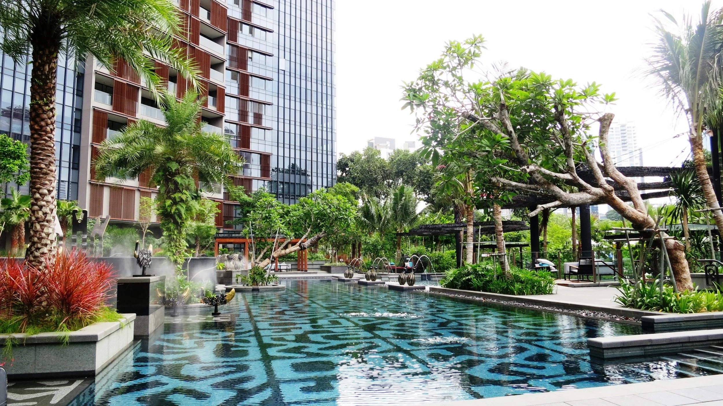 Urban pool area surrounded by tropical plants, trees, and a tall modern building with glass windows.
