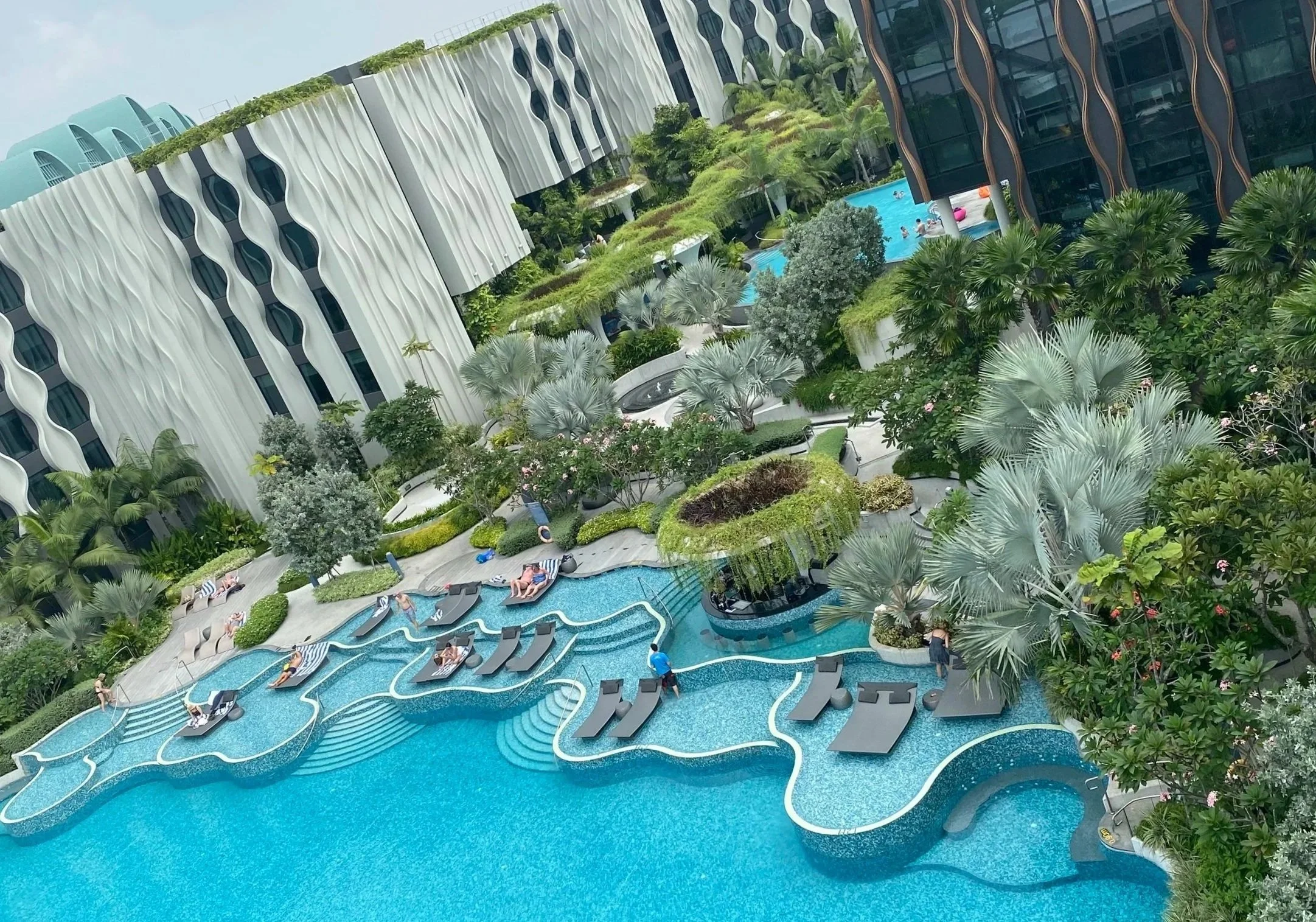 Aerial view of a luxury hotel pool with winding water slides, surrounded by lush greenery and modern buildings with unique architectural designs.