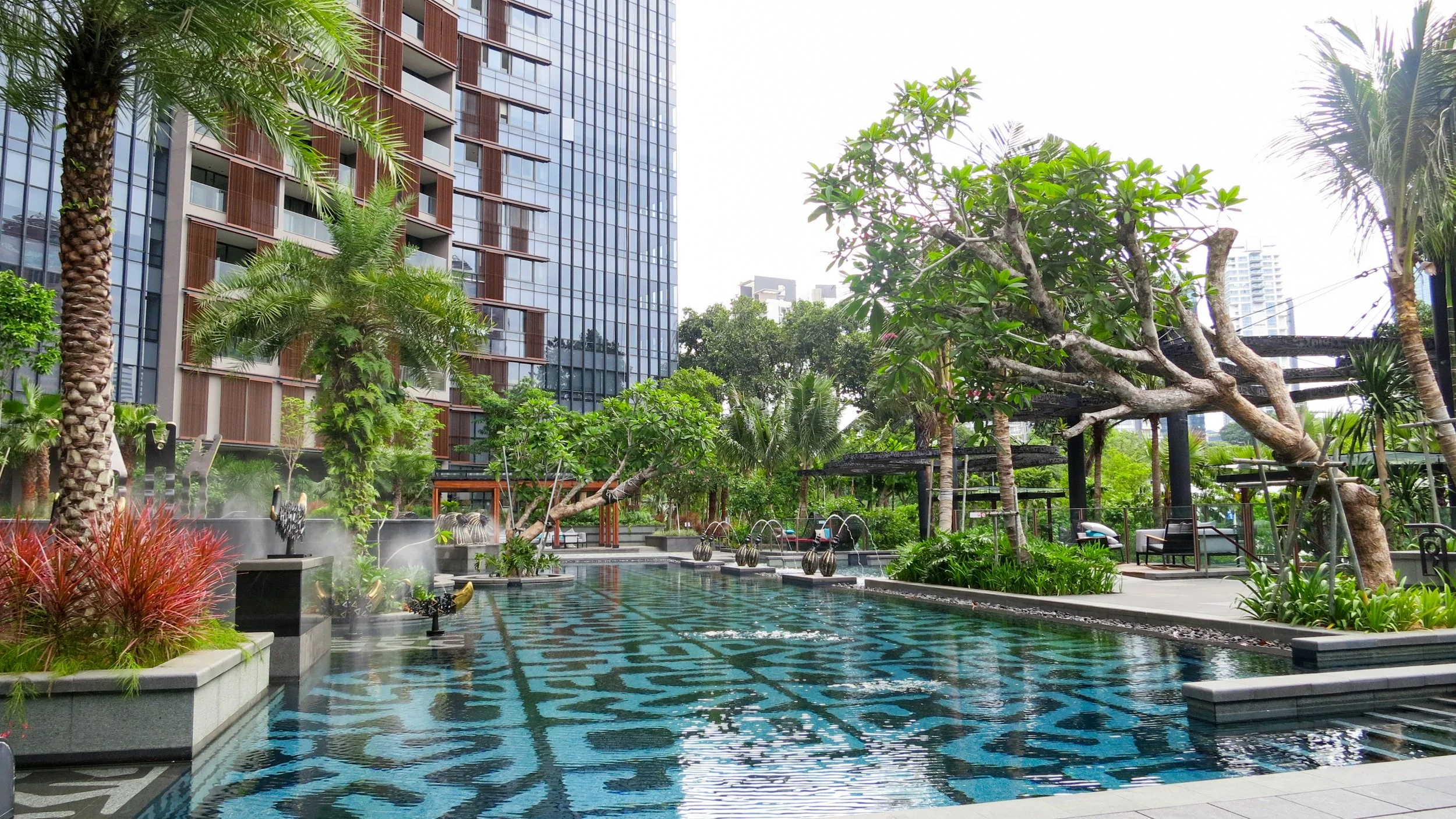 Urban outdoor swimming pool surrounded by lush green trees and plants, with a modern high-rise building in the background.
