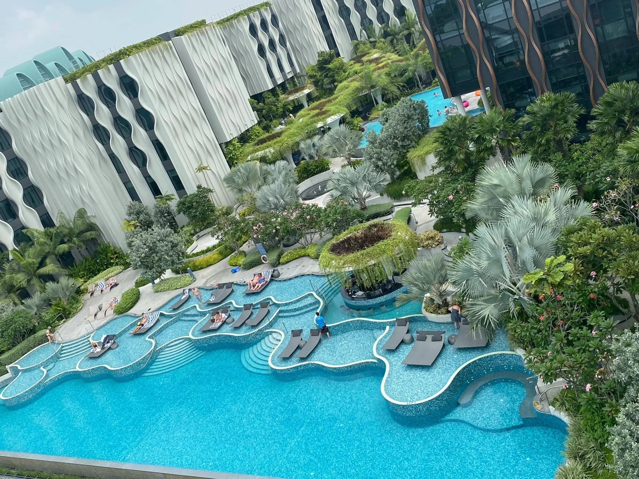 A swimming pool with a winding water slide surrounded by lush tropical plants and trees, with a modern high-rise building in the background.