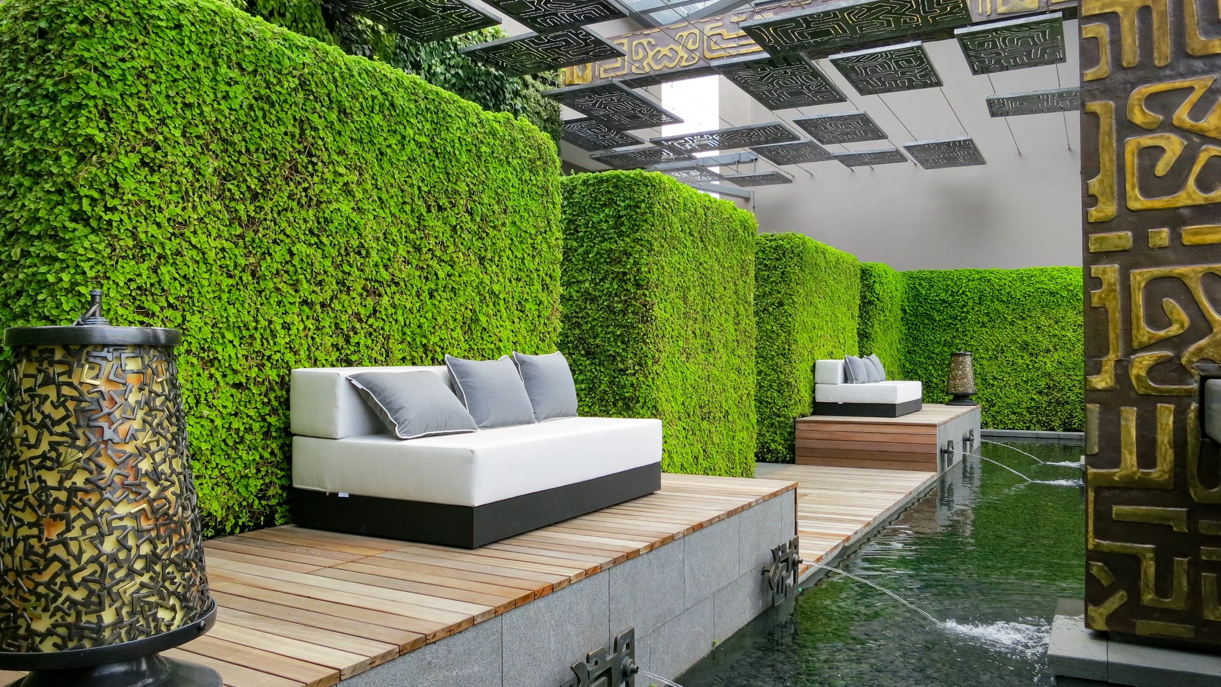 Outdoor seating area with white sofa and gray pillows, green hedge wall, decorative black and bronze metal lamp, water feature with small fountains, and metal art panels hanging overhead.