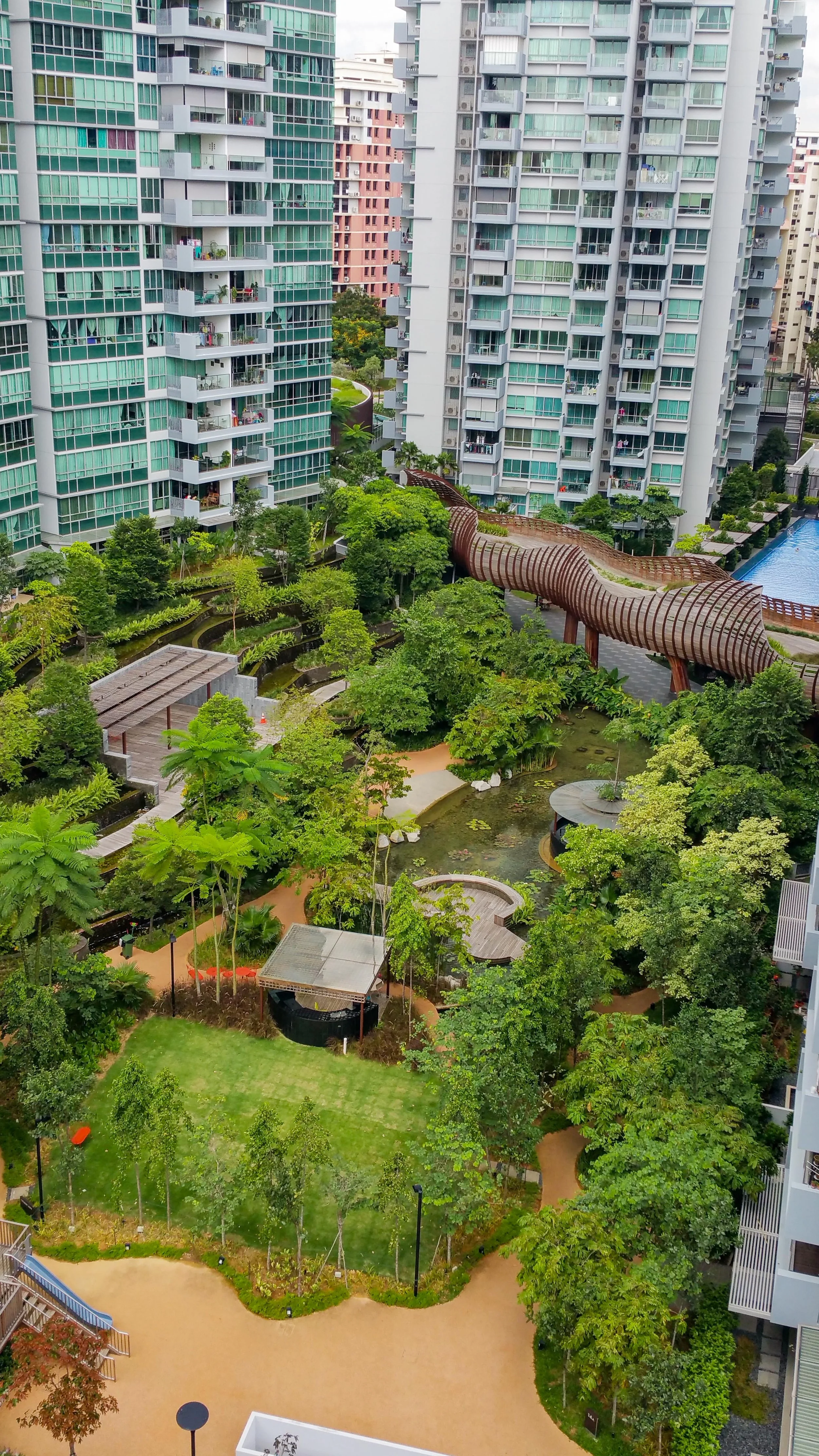 High-rise residential buildings surrounding a lush green garden with walking paths, trees, a pond, and a wooden observation walkway in an urban area.