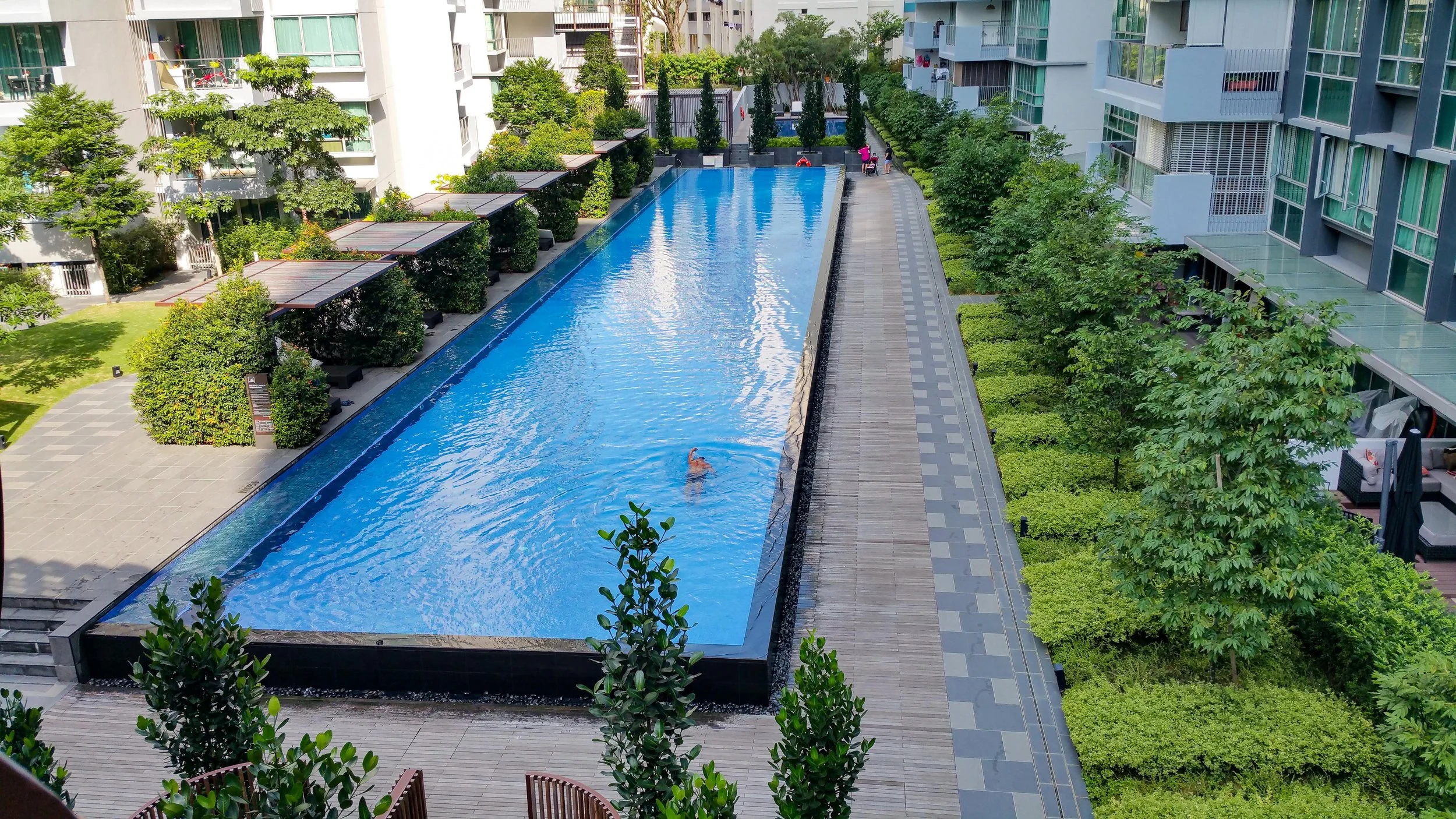 View of a swimming pool in a residential complex with green trees and shrubbery on either side, surrounded by modern apartment buildings with balconies and large glass windows. A person is swimming in the pool, and a few people are walking on the pathway nearby.