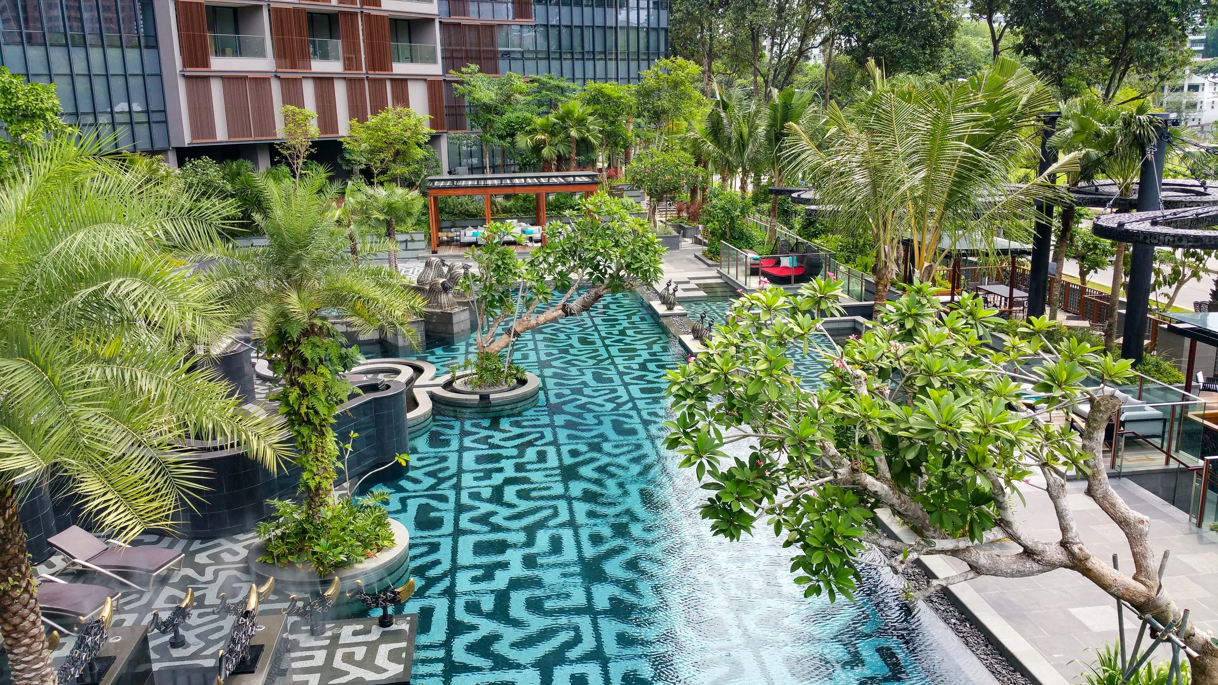 A luxurious outdoor swimming pool area with blue patterned tiles, surrounded by lush green trees and plants, with modern seating and architectural buildings in the background.
