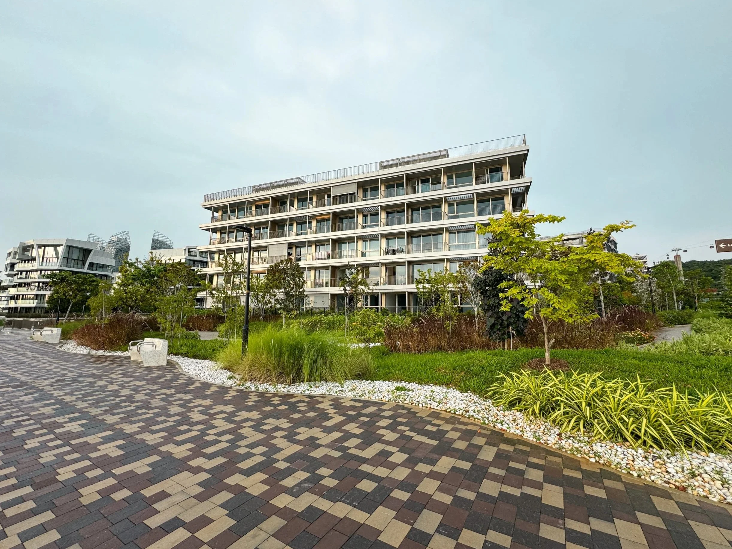 Modern multi-story apartment building with balconies, surrounded by landscaped greenery, trees, and a paved walkway.