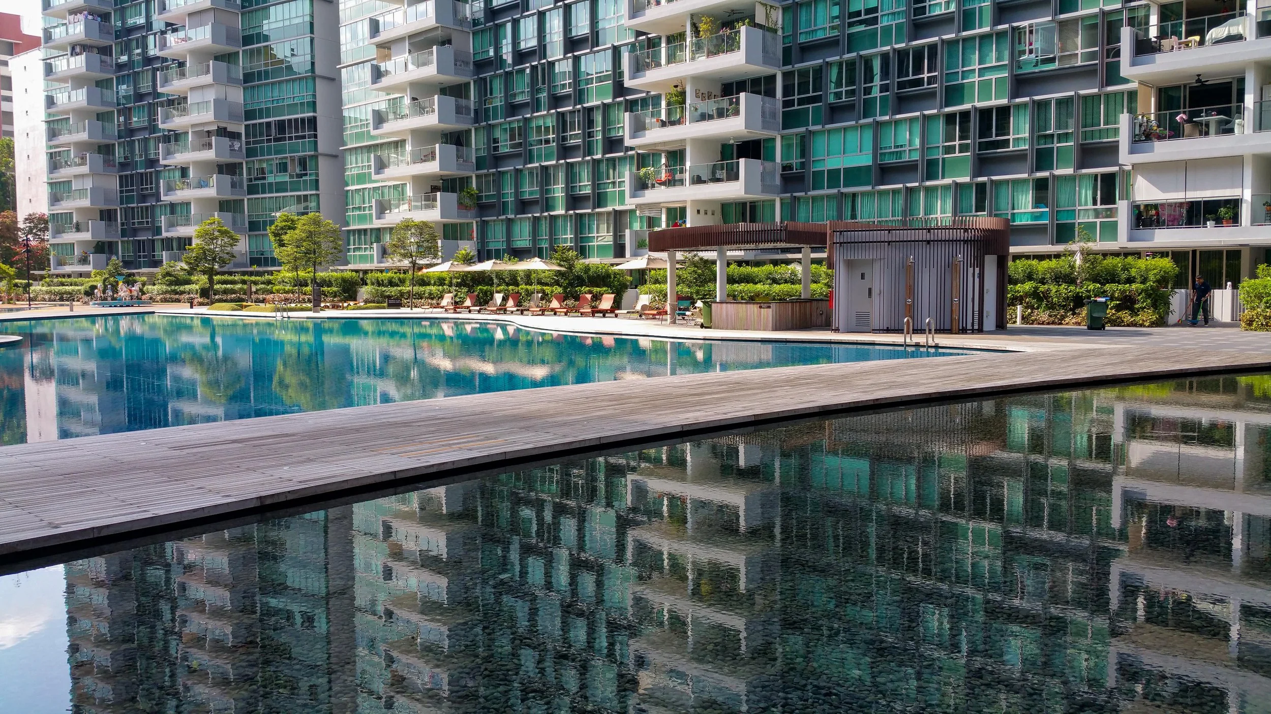 A modern residential apartment complex with balconies and glass windows reflects in a swimming pool. Lounge chairs and trees are around the pool area.