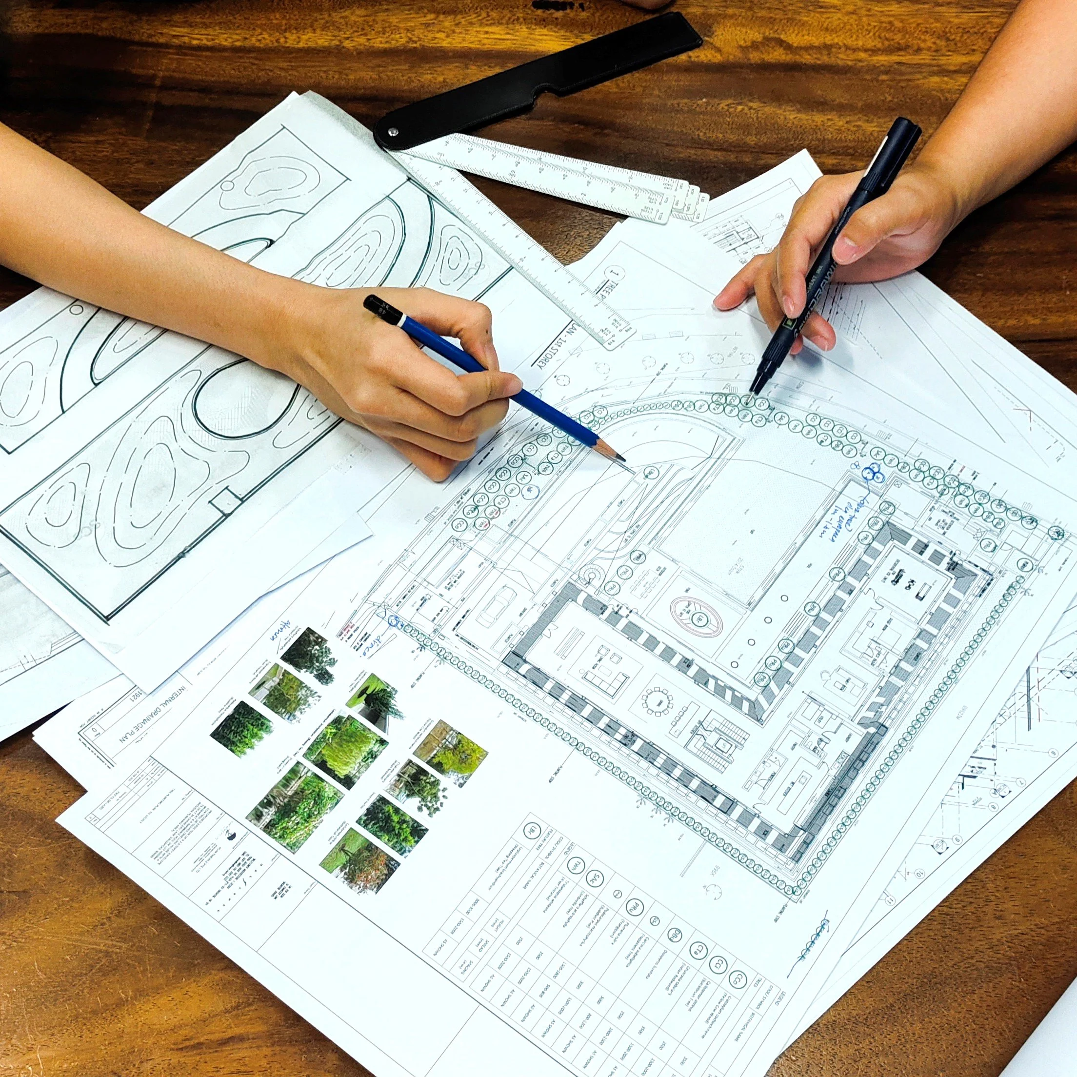 People working on landscape architecture plans with blueprints, a ruler, and a pen on a wooden surface.