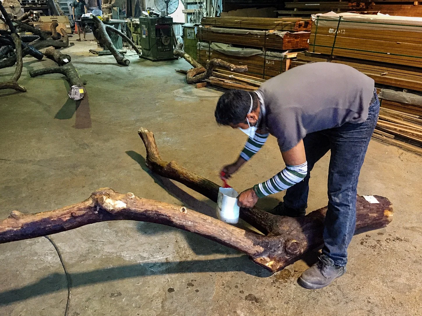 A man wearing a face mask, gray t-shirt, and jeans is bending over, applying a liquid from a plastic container onto a large wooden log with a brush in a woodworking workshop. Several logs and wooden planks are stacked in the background.