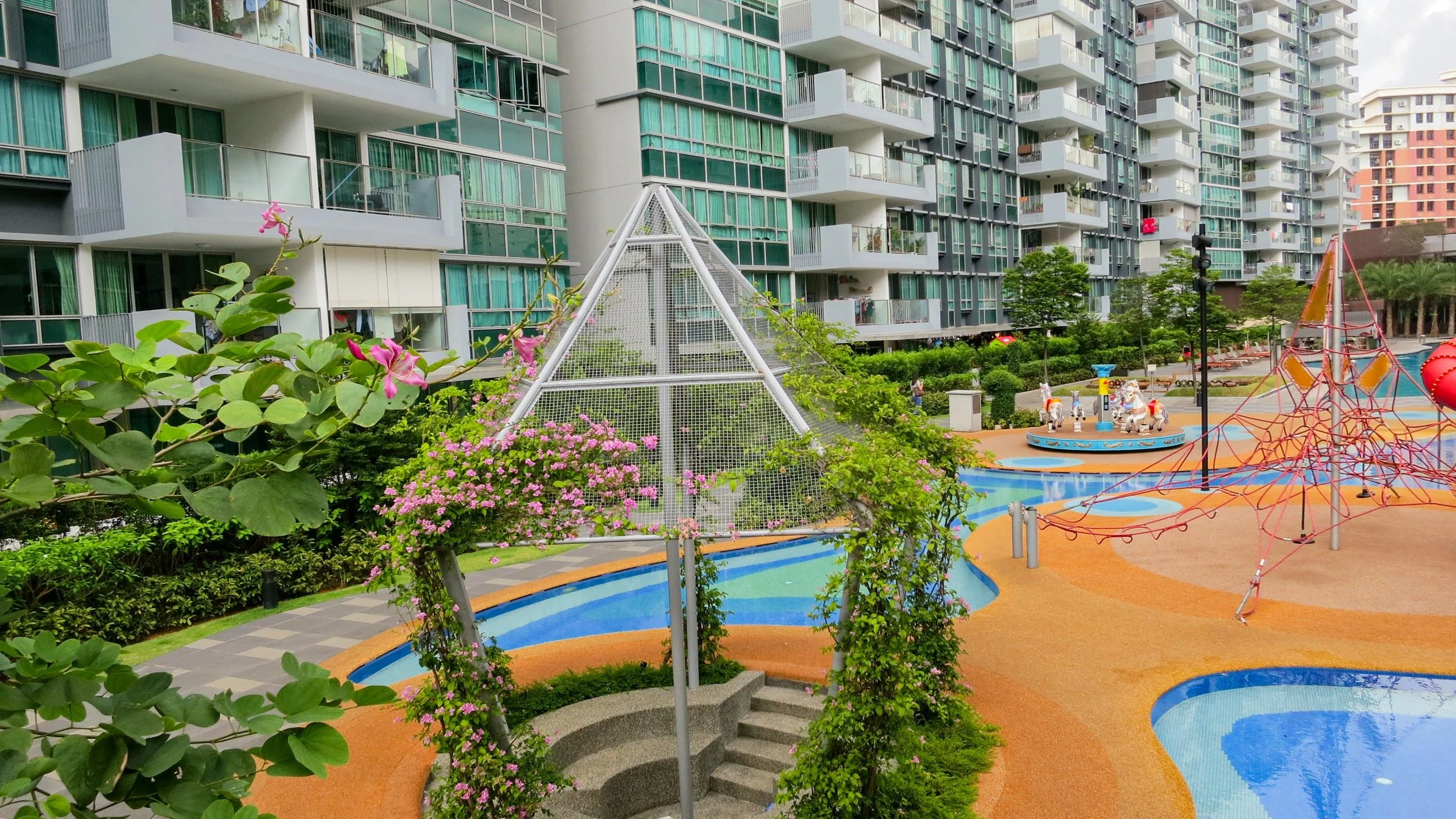 View of a colorful children's playground with a carousel, climbing structures, and a water feature, surrounded by greenery and high-rise apartment buildings.