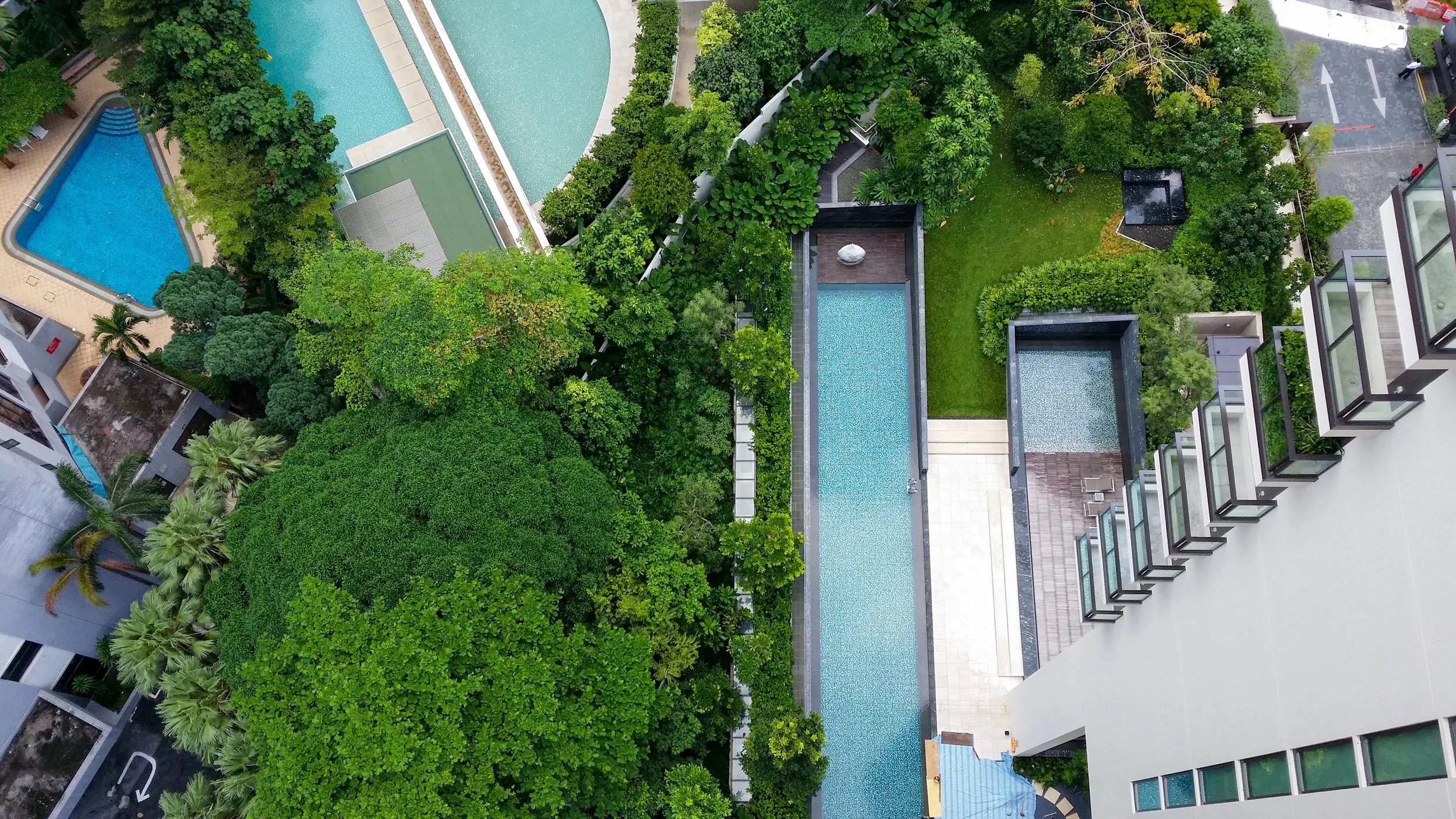 An aerial view of a modern apartment complex with outdoor pools, surrounded by lush green trees and landscaped gardens.