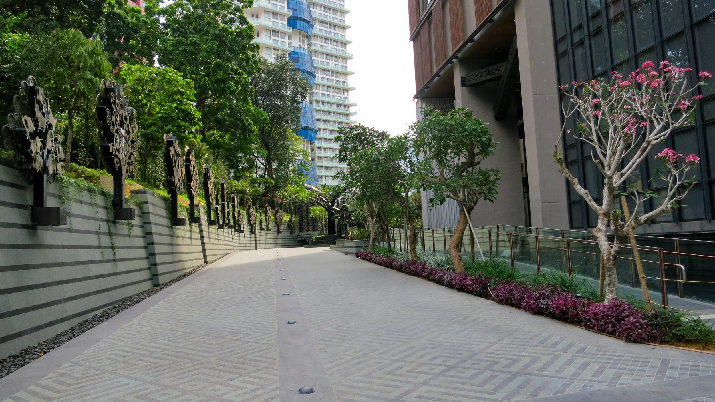 Urban outdoor walkway flanked by modern buildings, with trees and pink flowering plants on one side and decorative black metal sculptures along a striped concrete wall on the other