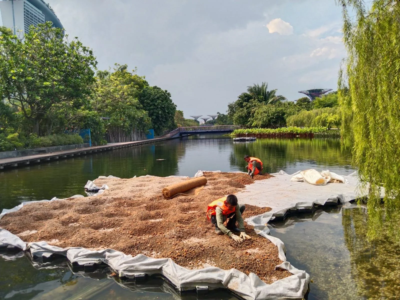 Individuals working on a construction project on a floating platform in a river, surrounded by greenery and modern buildings in the background.