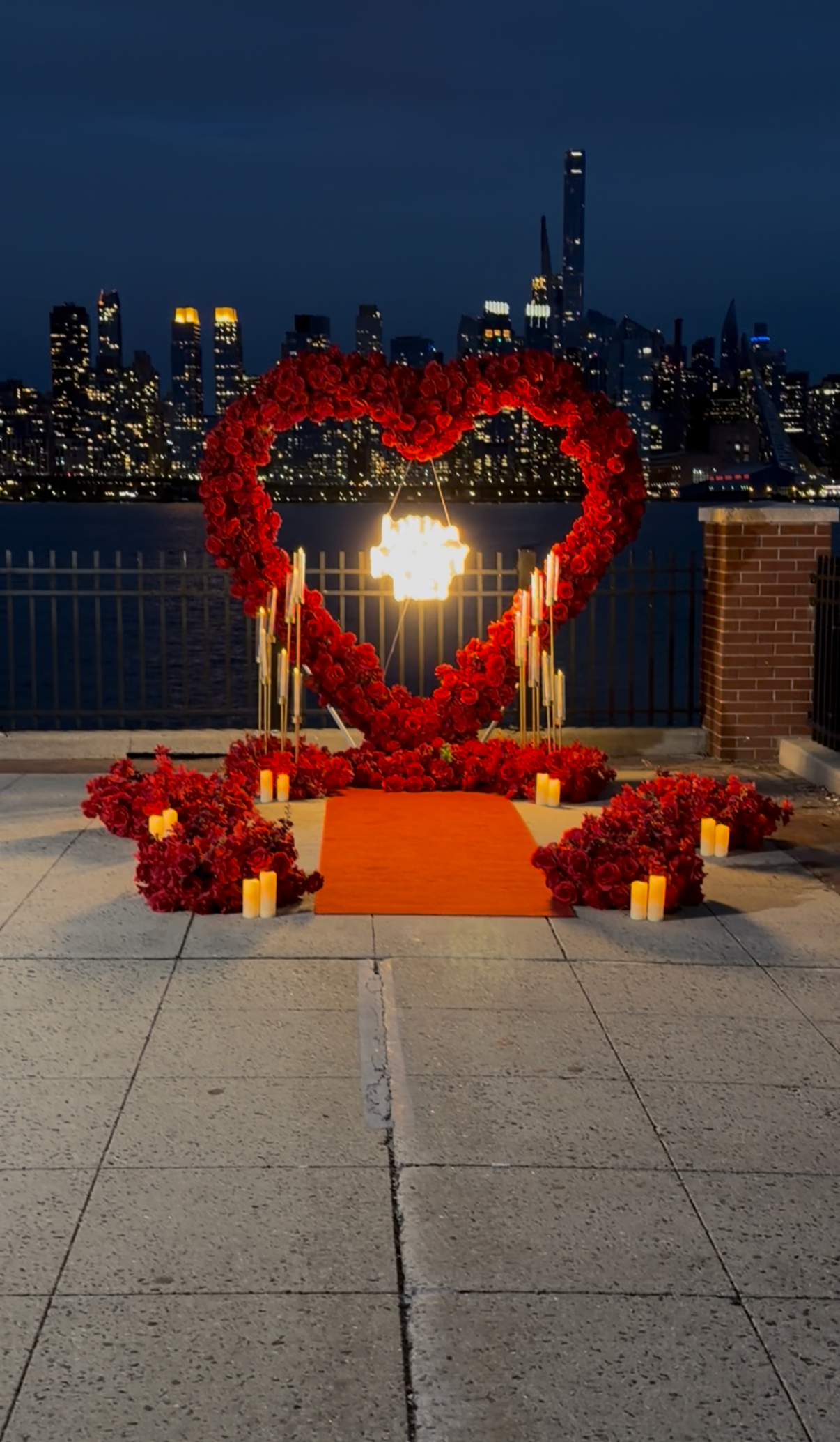 A romantic setup on a rooftop featuring a large heart-shaped arch made of red roses, with a candle chandelier hanging inside the heart. The scene is decorated with candles and flower arrangements around a red carpet, overlooking a city skyline at night.