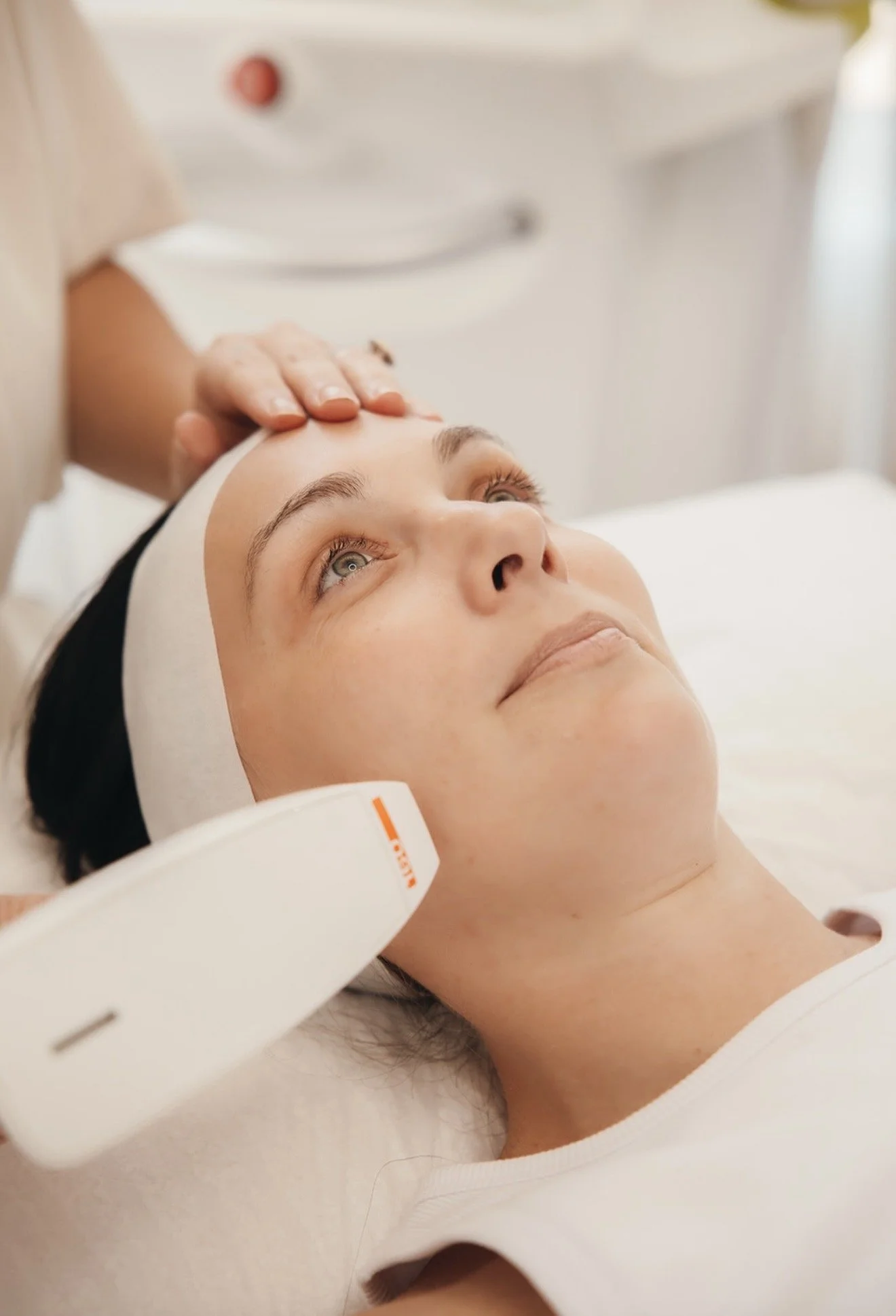 A woman lying on a treatment bed receiving a facial massage, wearing a headband, with a white medical device or tool near her neck.