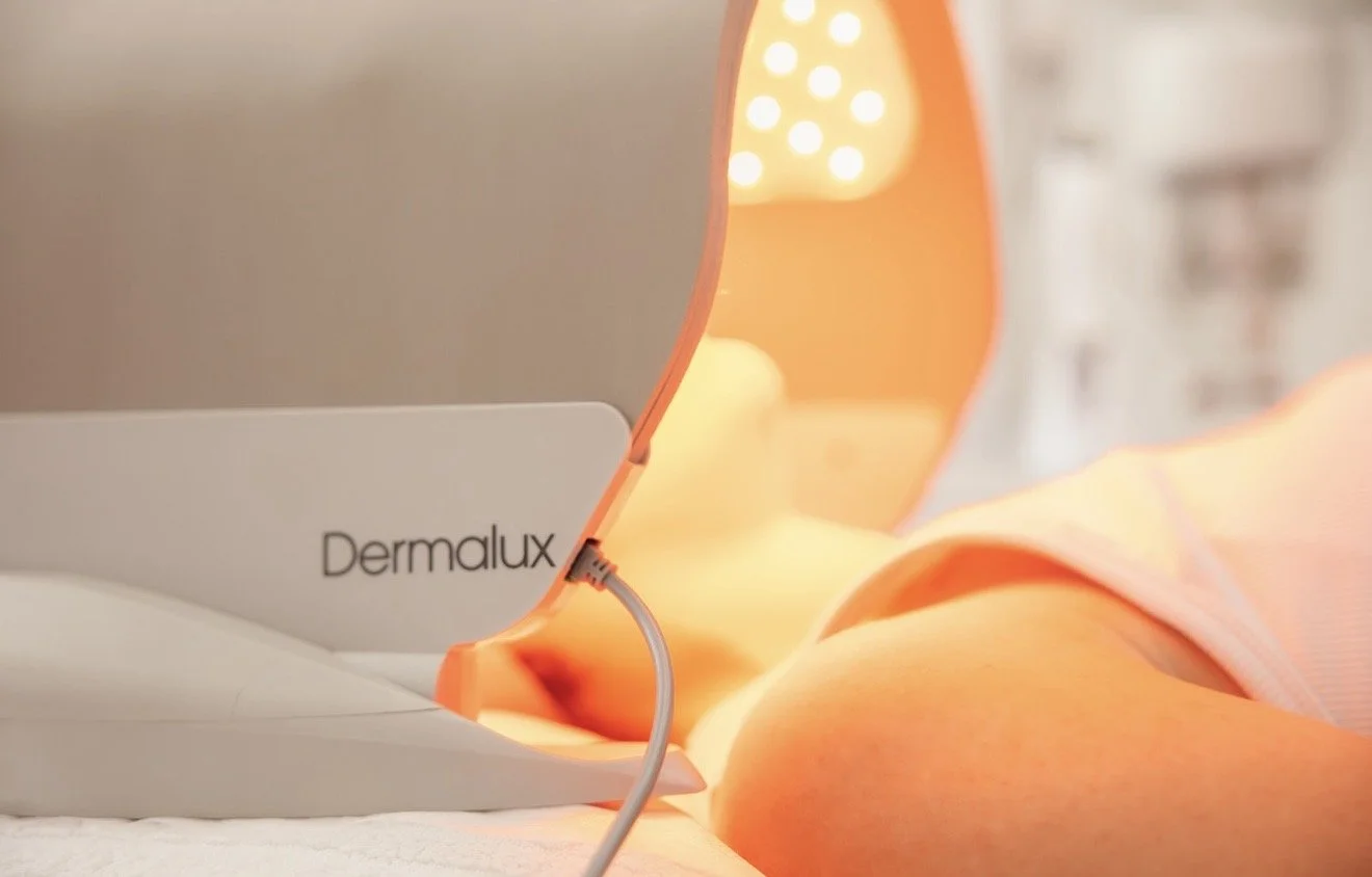 Close-up of a dermatology LED light therapy device labeled 'Dermalux' emitting warm light, with a person's face partially visible during treatment.