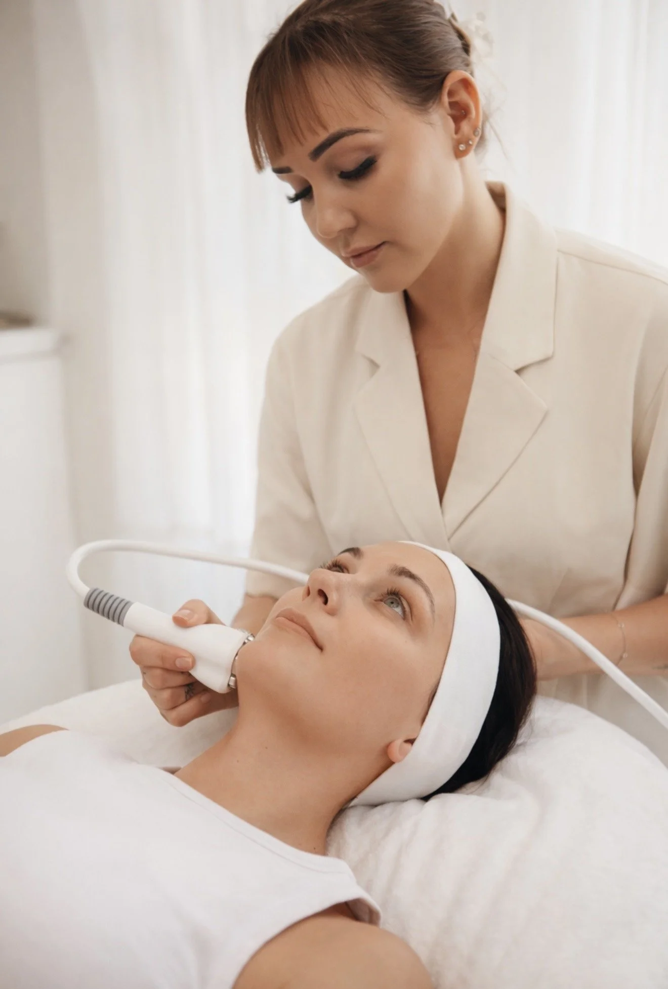 A woman receiving a facial treatment with a handheld device from a skincare professional in a spa or clinic setting.