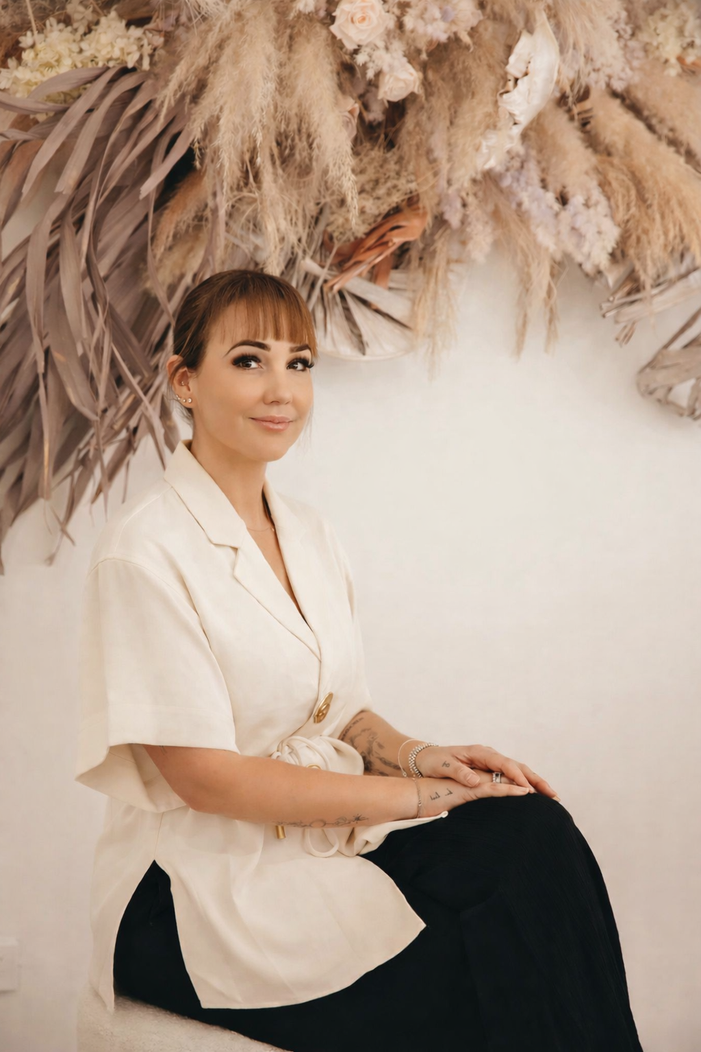 A woman with short brown hair and bangs sitting against a white wall with a large arrangement of dried pampas grass and foliage behind her.