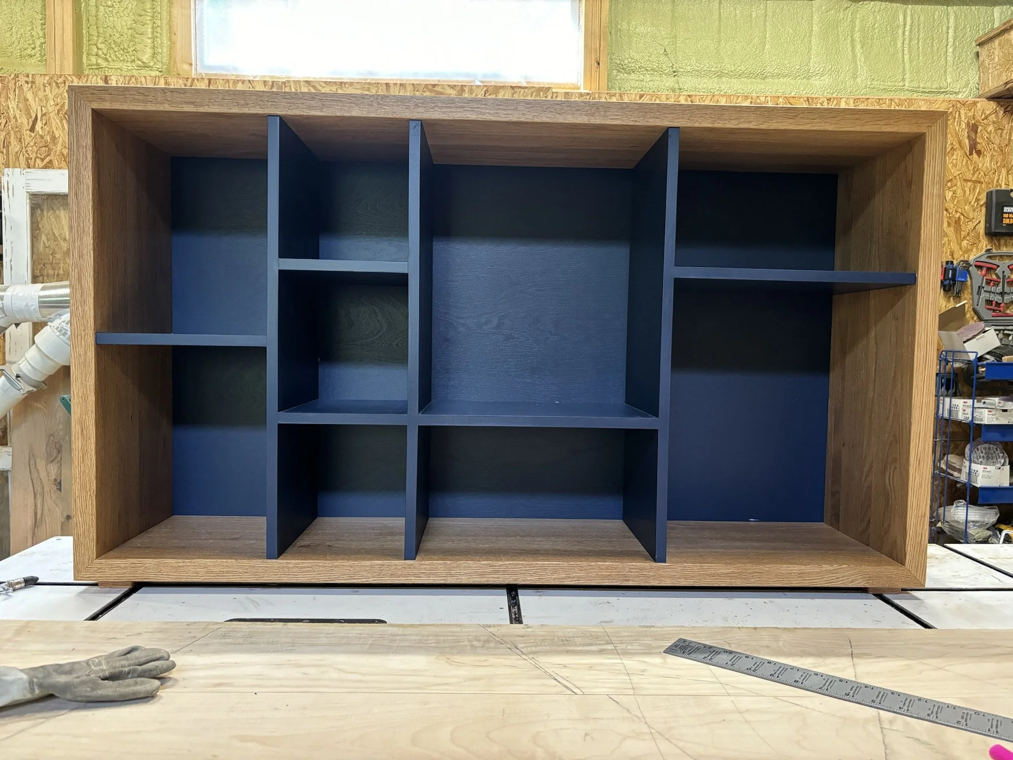 Wooden shelving unit with multiple compartments painted dark blue, situated on a workbench in a woodworking shop.