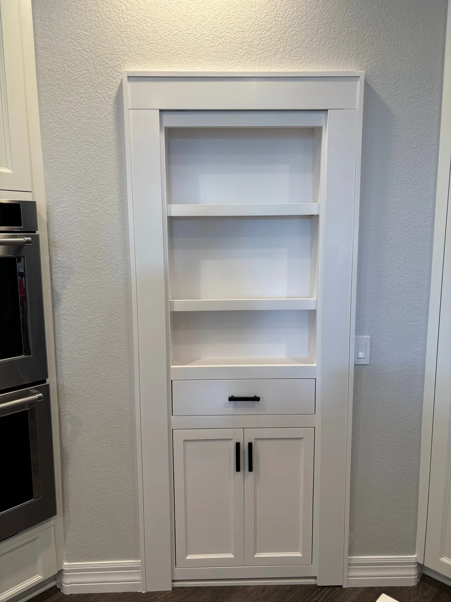 White built-in kitchen cabinet with open shelves, a drawer, and closed cabinet doors, situated against a light-colored textured wall.