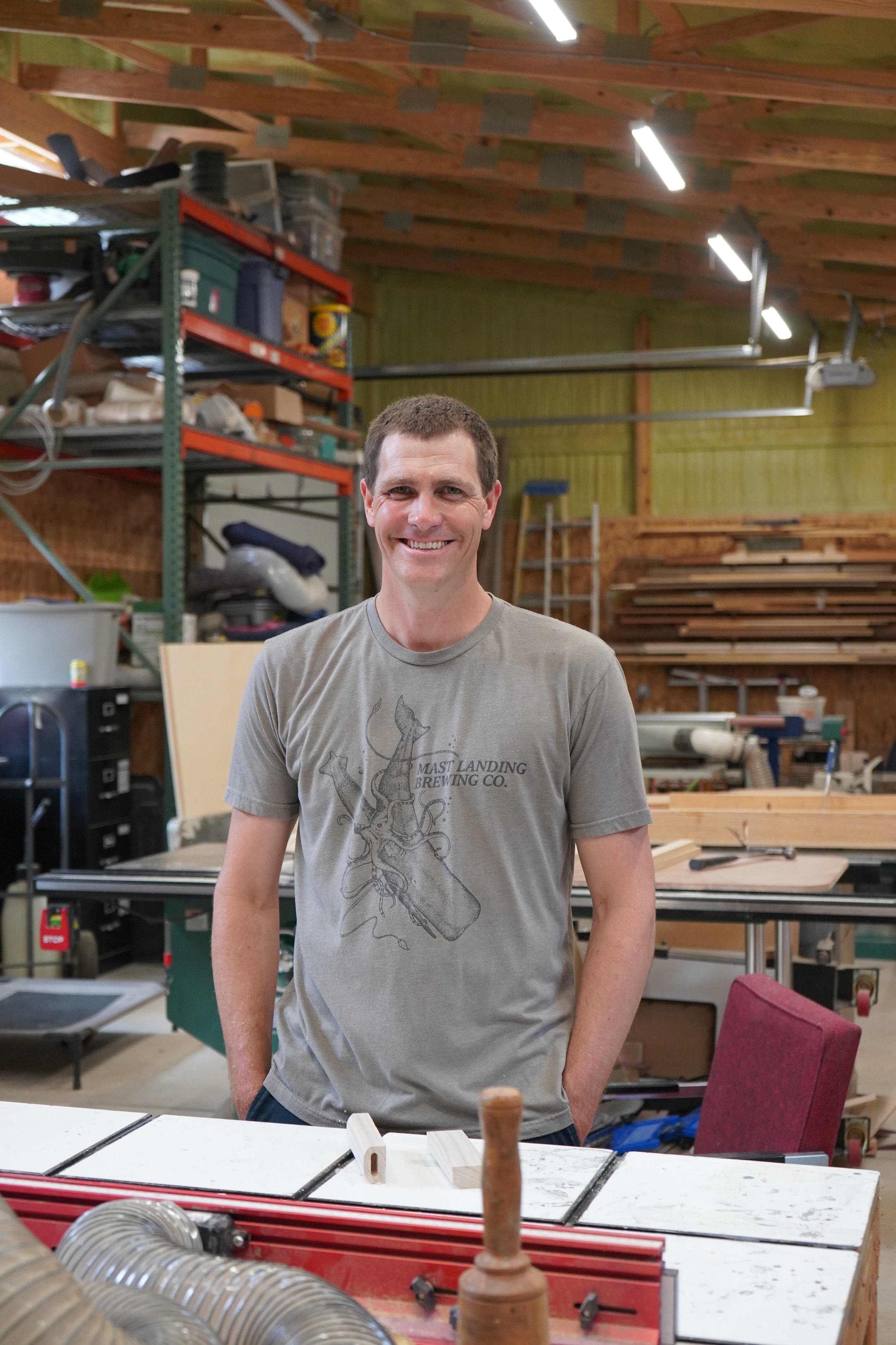 A smiling man standing in a woodworking workshop with tools and wood pieces in the background.