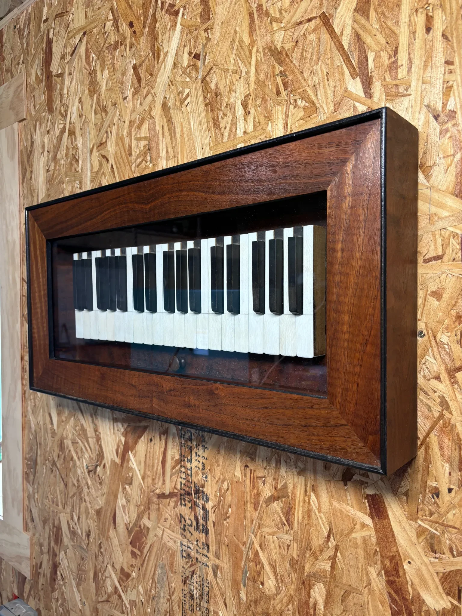 A miniature piano with black and white keys, encased in a wooden frame with a glass cover, placed on a wooden surface.