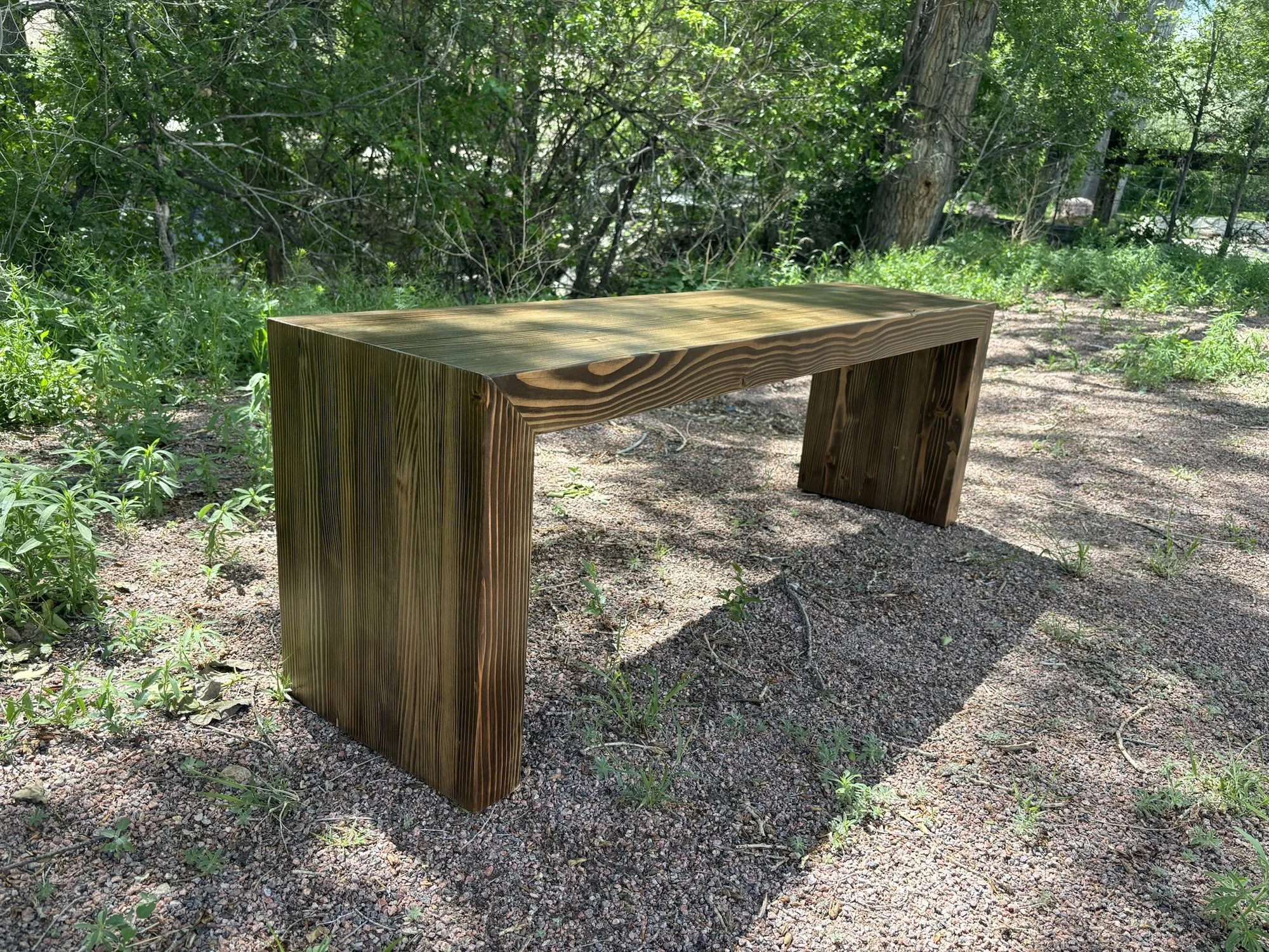 A wooden bench on a dirt surface surrounded by greenery and trees in an outdoor setting.