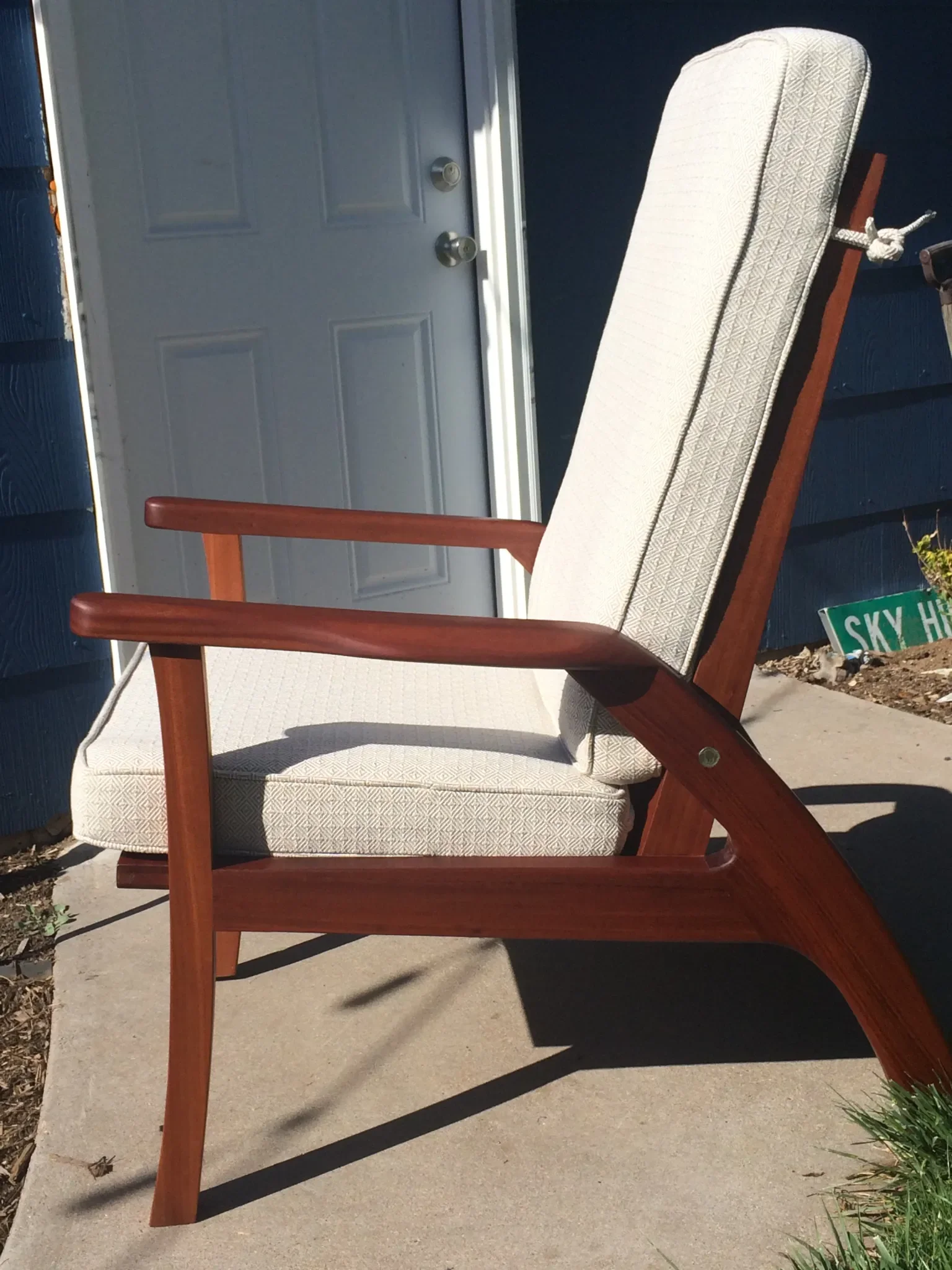A wooden outdoor chair with a beige patterned cushion, placed on a concrete sidewalk in front of a blue house with a gray door.
