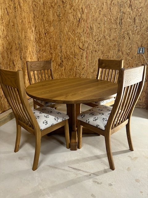A wooden round dining table with four matching chairs, two with upholstered seats featuring a paw print pattern, in a room with plywood walls and beige flooring.
