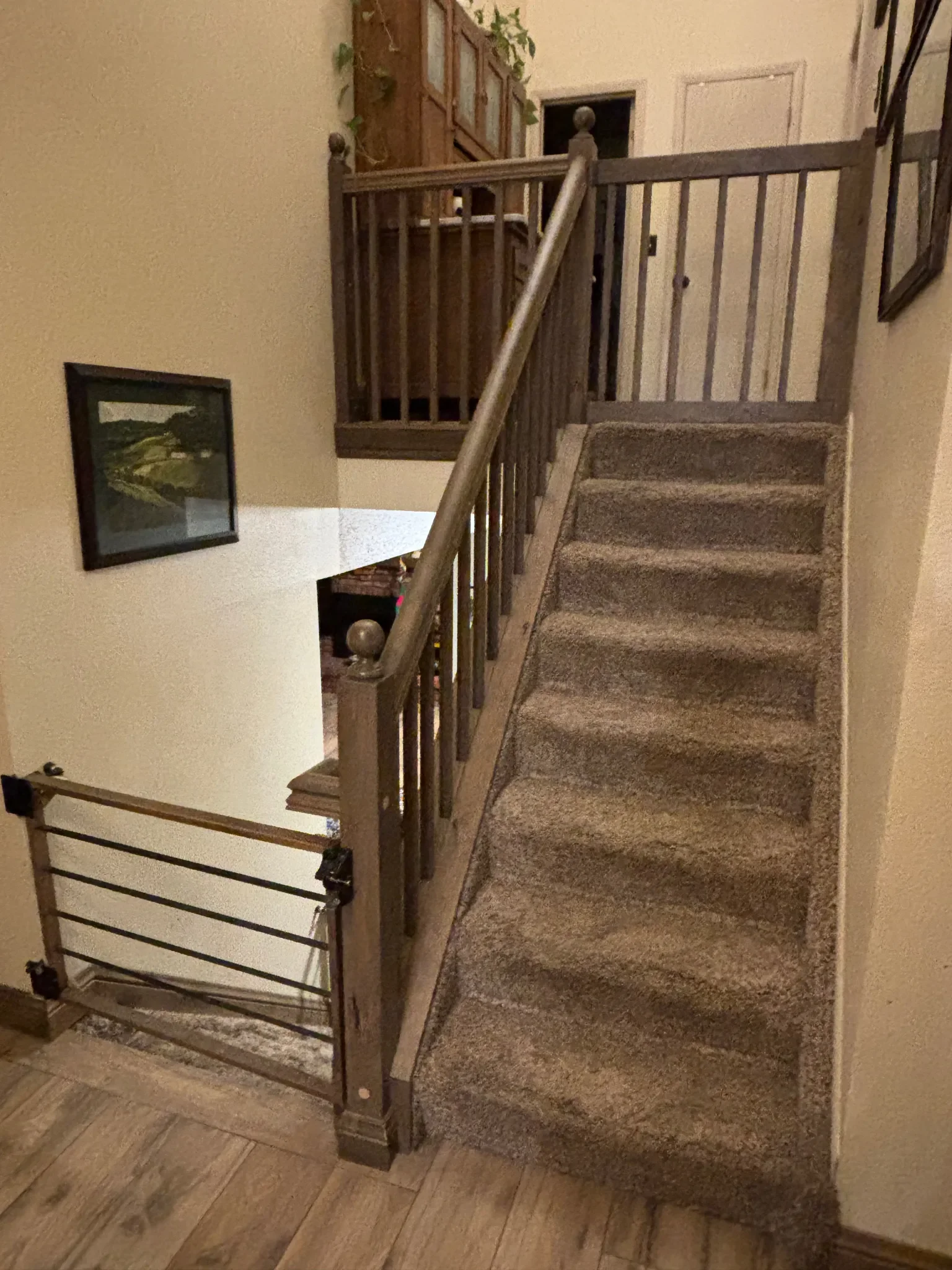 Carpeted staircase with a wooden banister leading to an upstairs area in a house, with framed pictures and a staircase gate at the bottom.