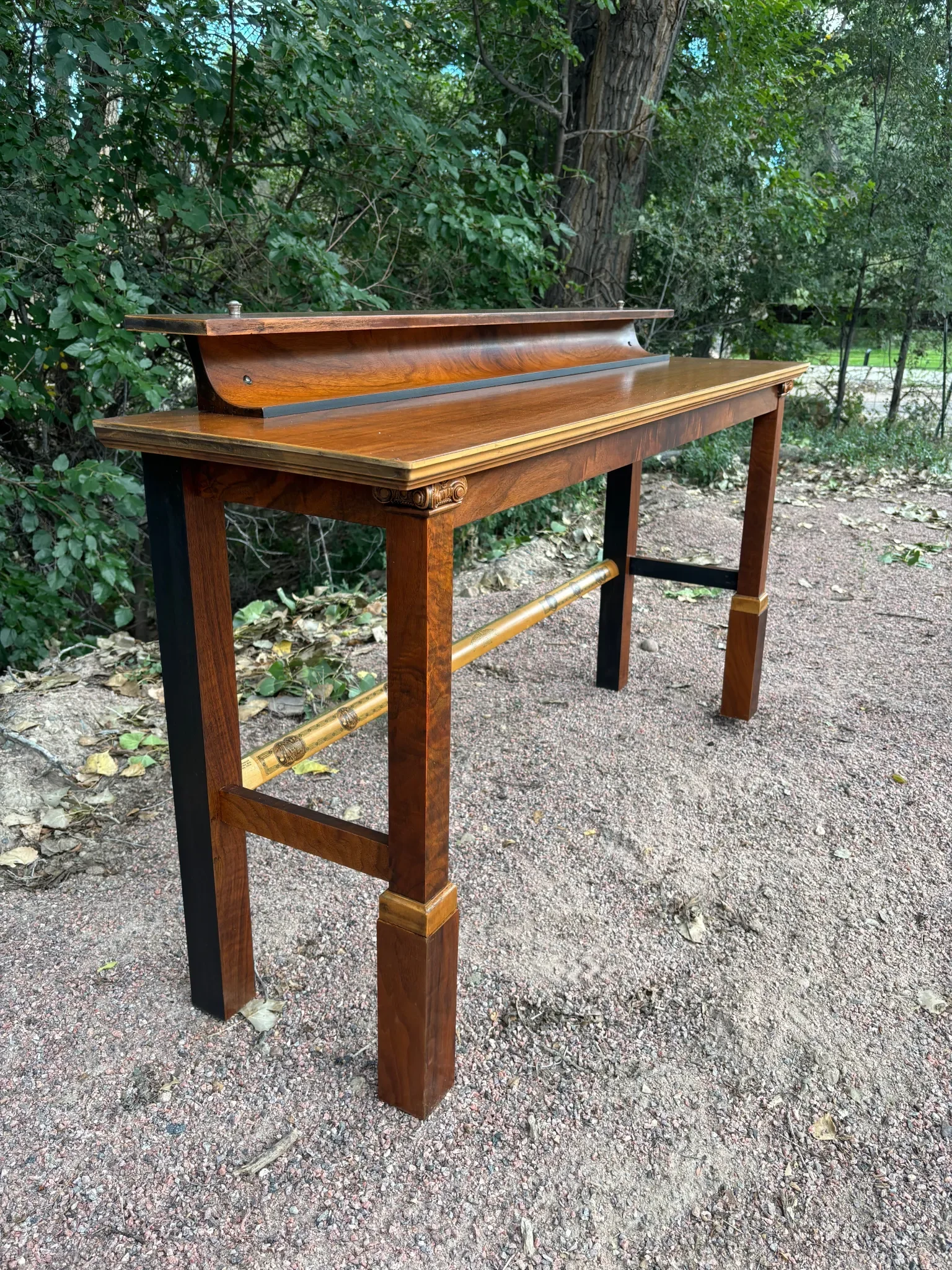 A vintage wooden piano with a polished finish, featuring decorative, carved column details on the legs, outdoors on a gravel path surrounded by trees and greenery.