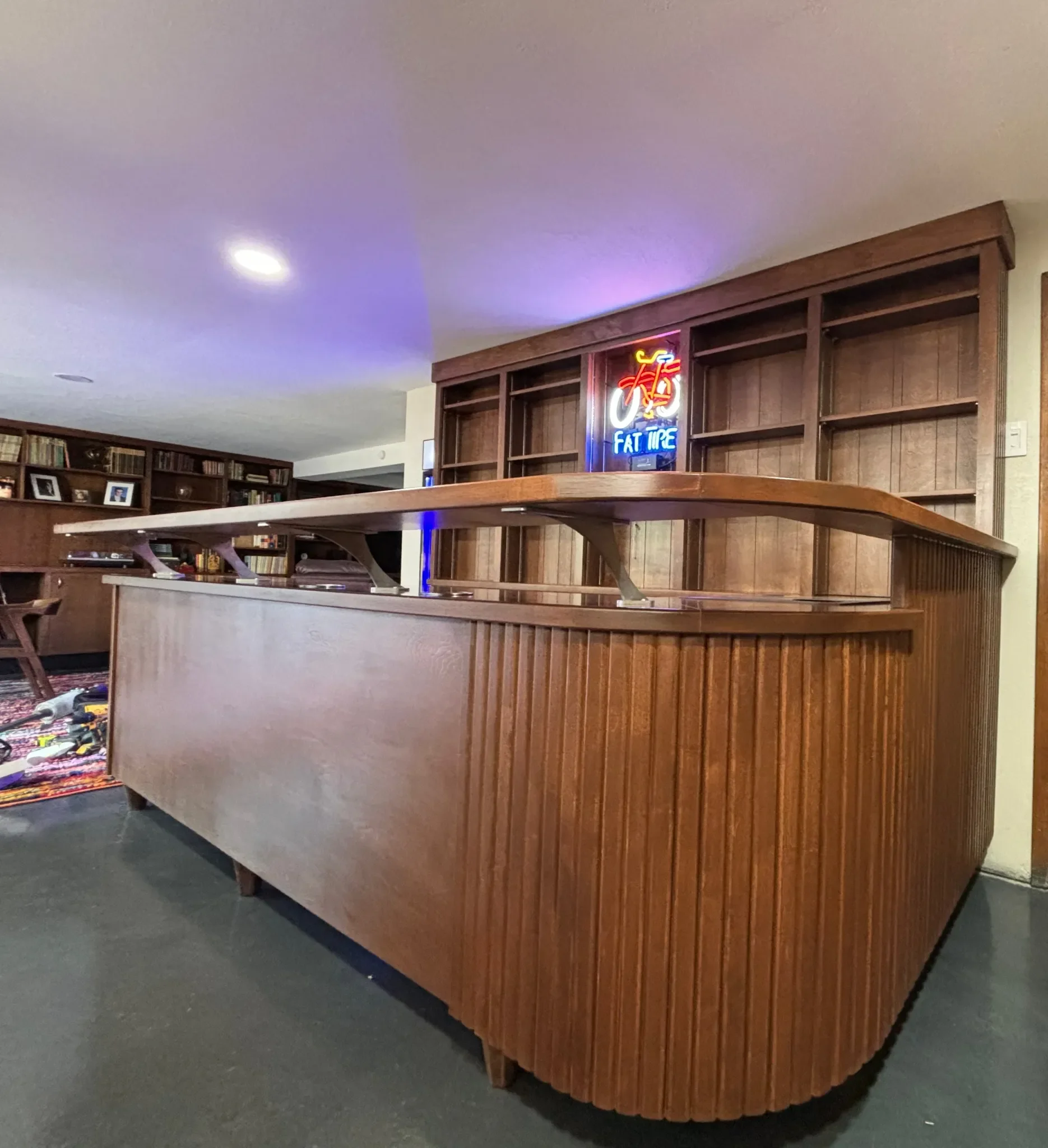 A wooden bar counter with a curved front and a higher shelf on top, in a room with bookshelves and a neon sign featuring a bicycle and the words "Fat Tire".
