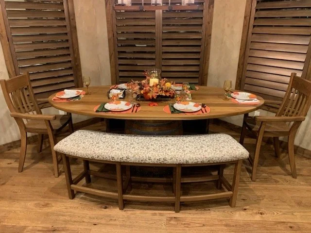 A wooden dining table set for four with plates, napkins, and glasses, and a floral centerpiece, with a cushioned bench and wooden chairs, in a room with wooden walls and flooring.