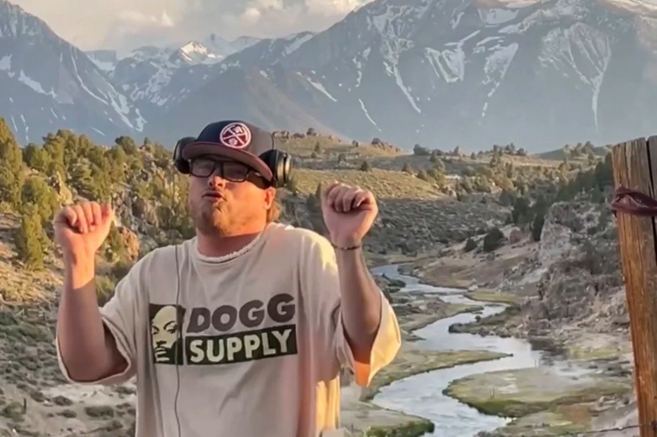 A man with red hair, beard, glasses, and headphones dancing outdoors in front of a mountainous landscape with a river, trees, and snow-capped peaks.