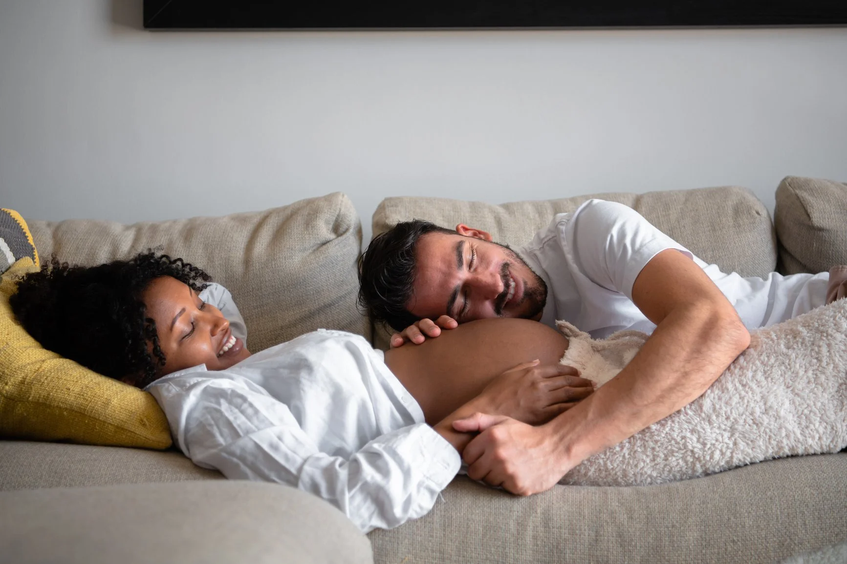 Pregnant couple cuddling on a couch, smiling as they share a tender moment together.