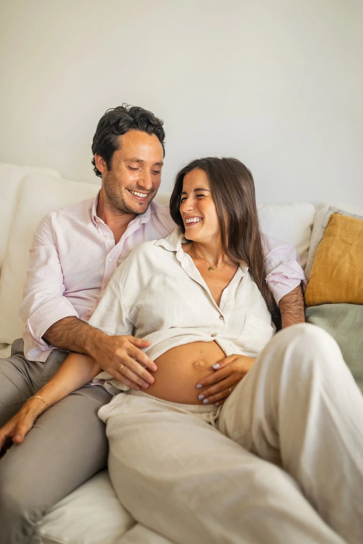 Pregnant couple sitting on couch and cuddling, while their hands rest on her belly.
