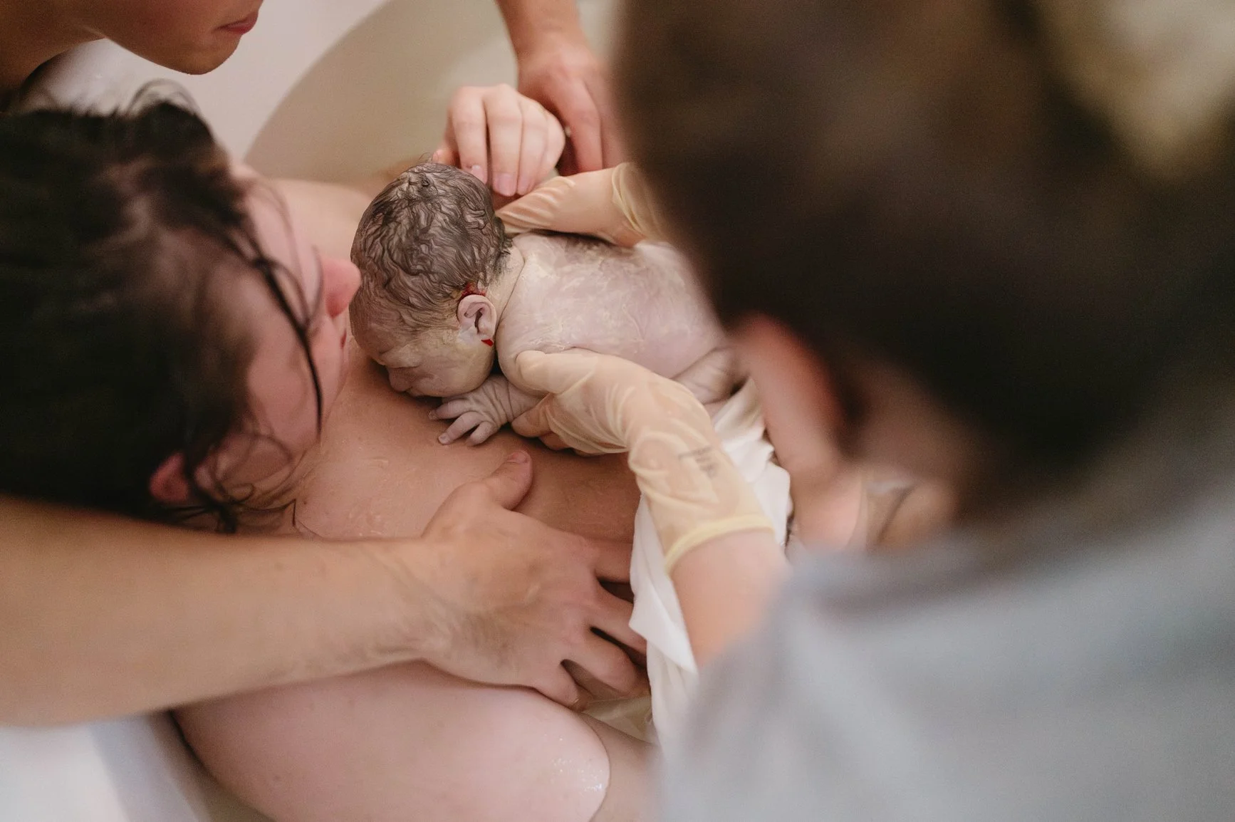 Woman lies in tub with her newborn against her chest while some people outside of the tub support her and the baby.