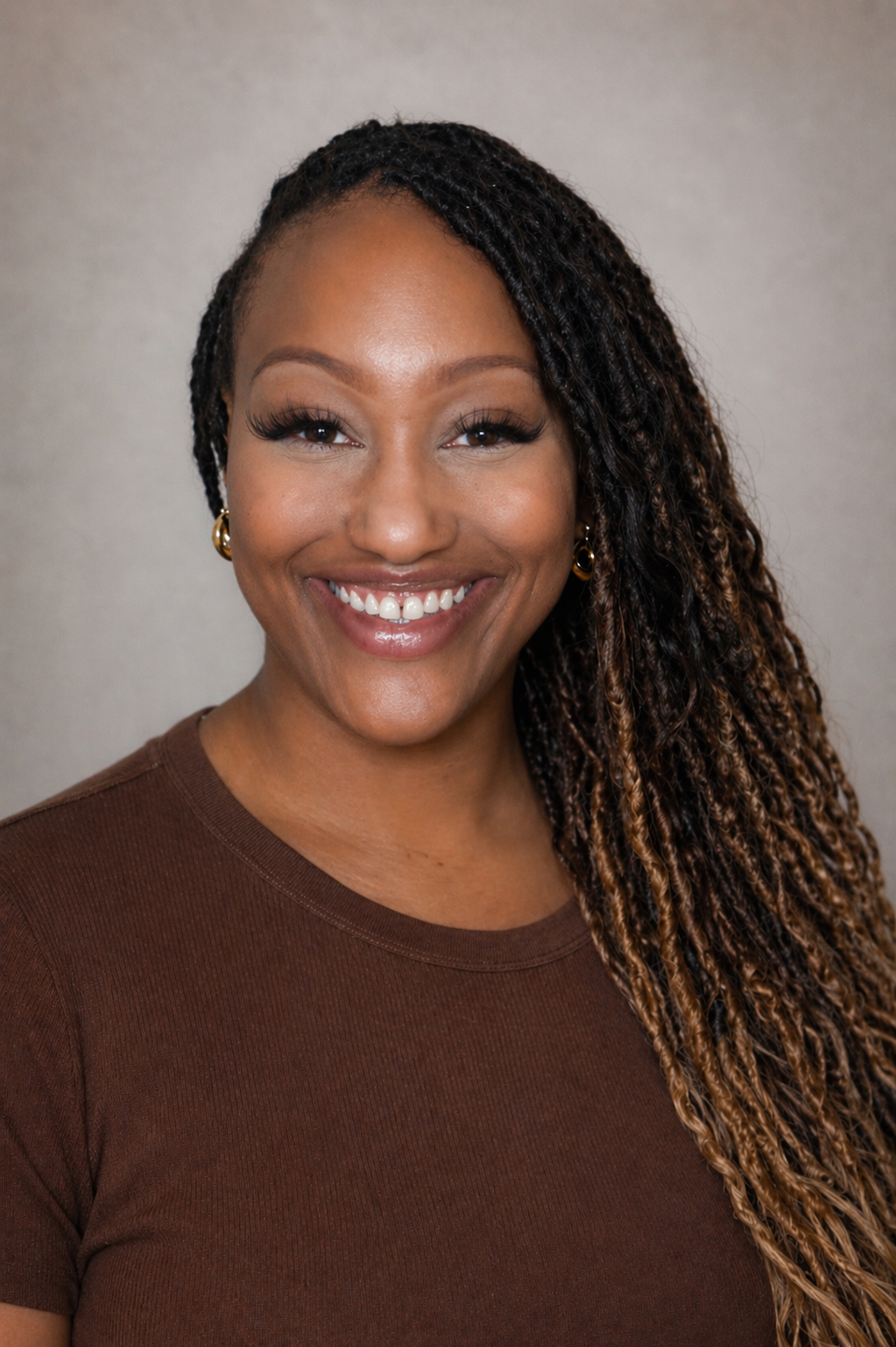 A woman with locs and hoop earrings smiles broadly, wearing a brown t-shirt against a neutral background.