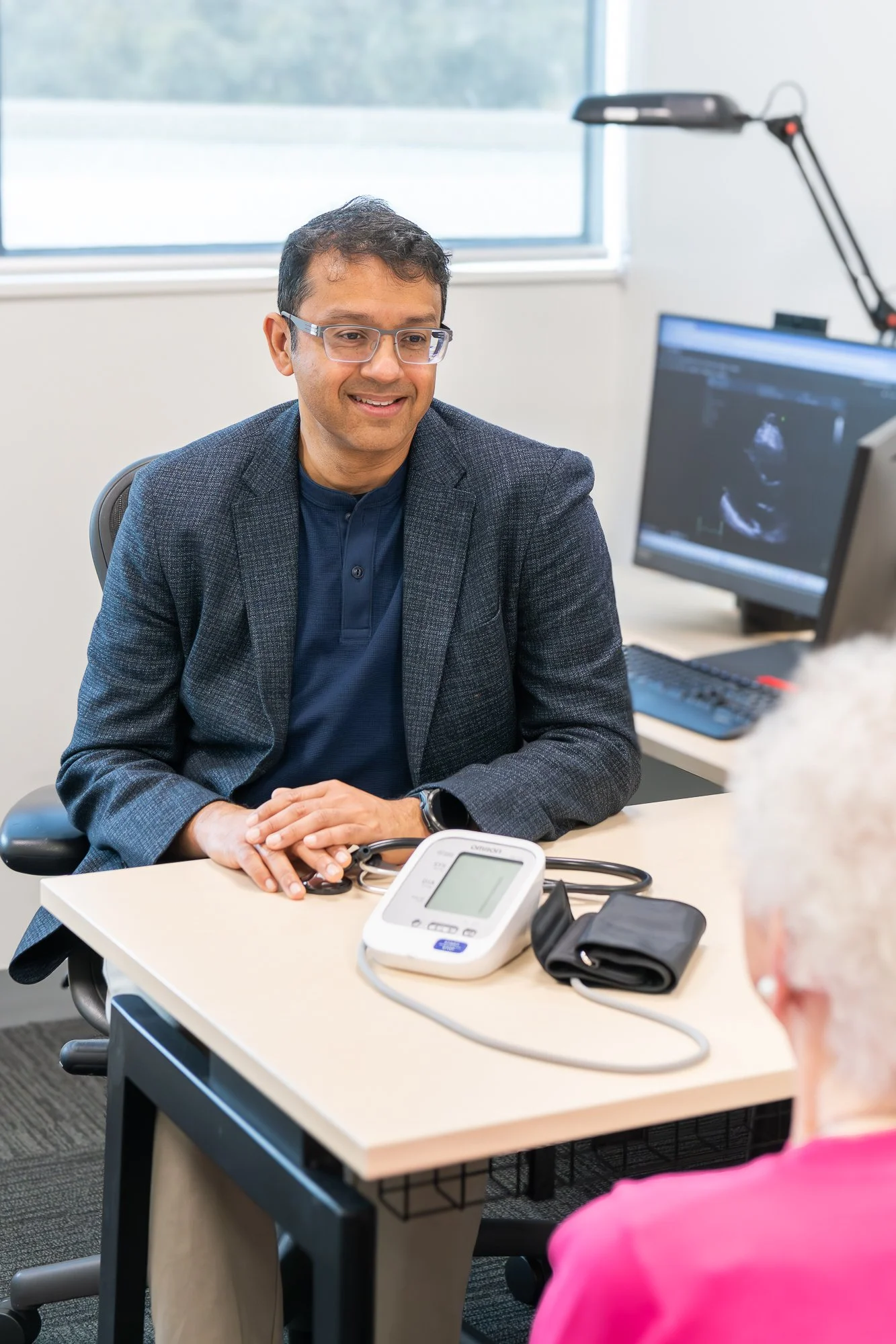 A male doctor wearing glasses and a dark blazer is smiling and sitting at a desk with medical equipment, including a blood pressure monitor and stethoscope, in a consultation with a patient at a medical office.