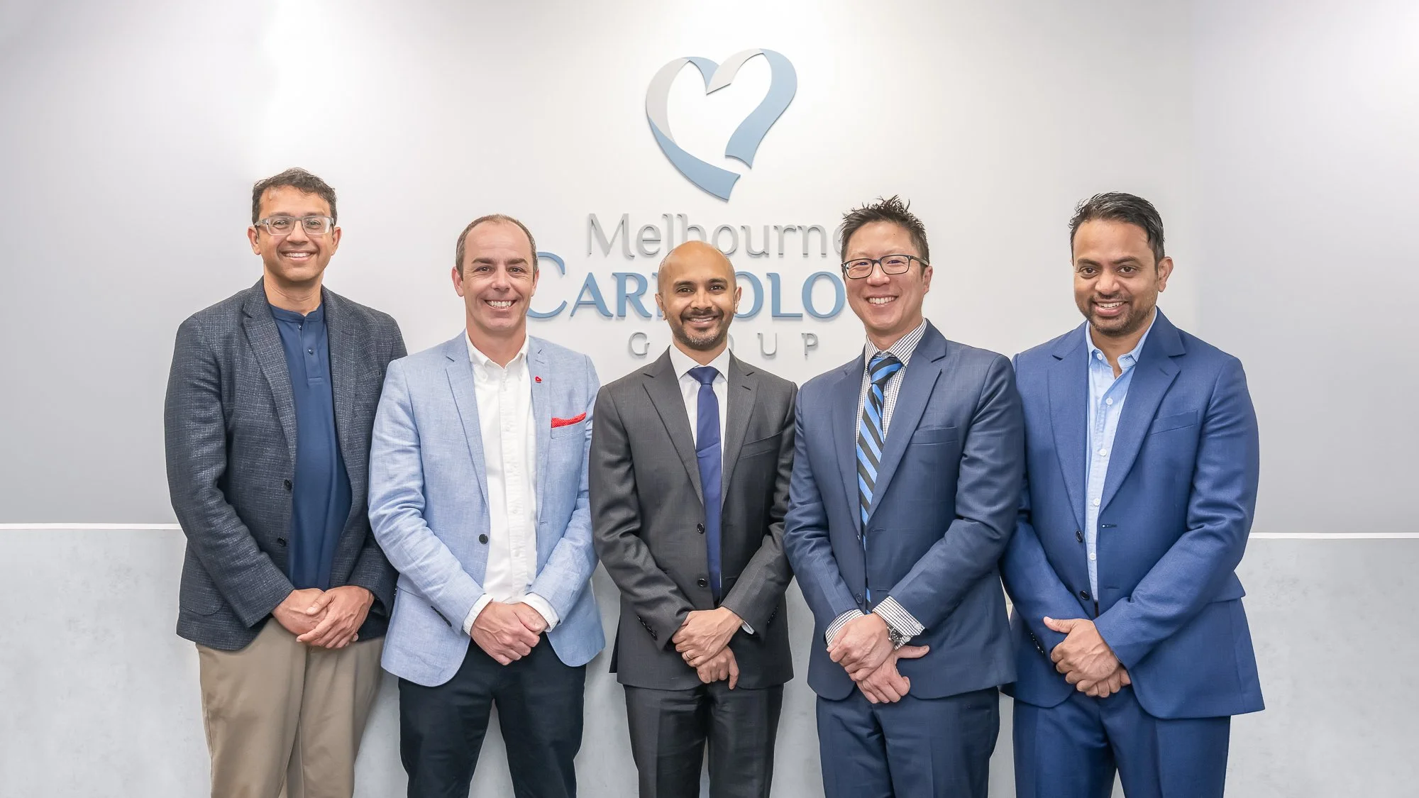 Group of five diverse men in business suits standing in front of a wall with a logo for Melbourne Cardiology Group, smiling for the photo.