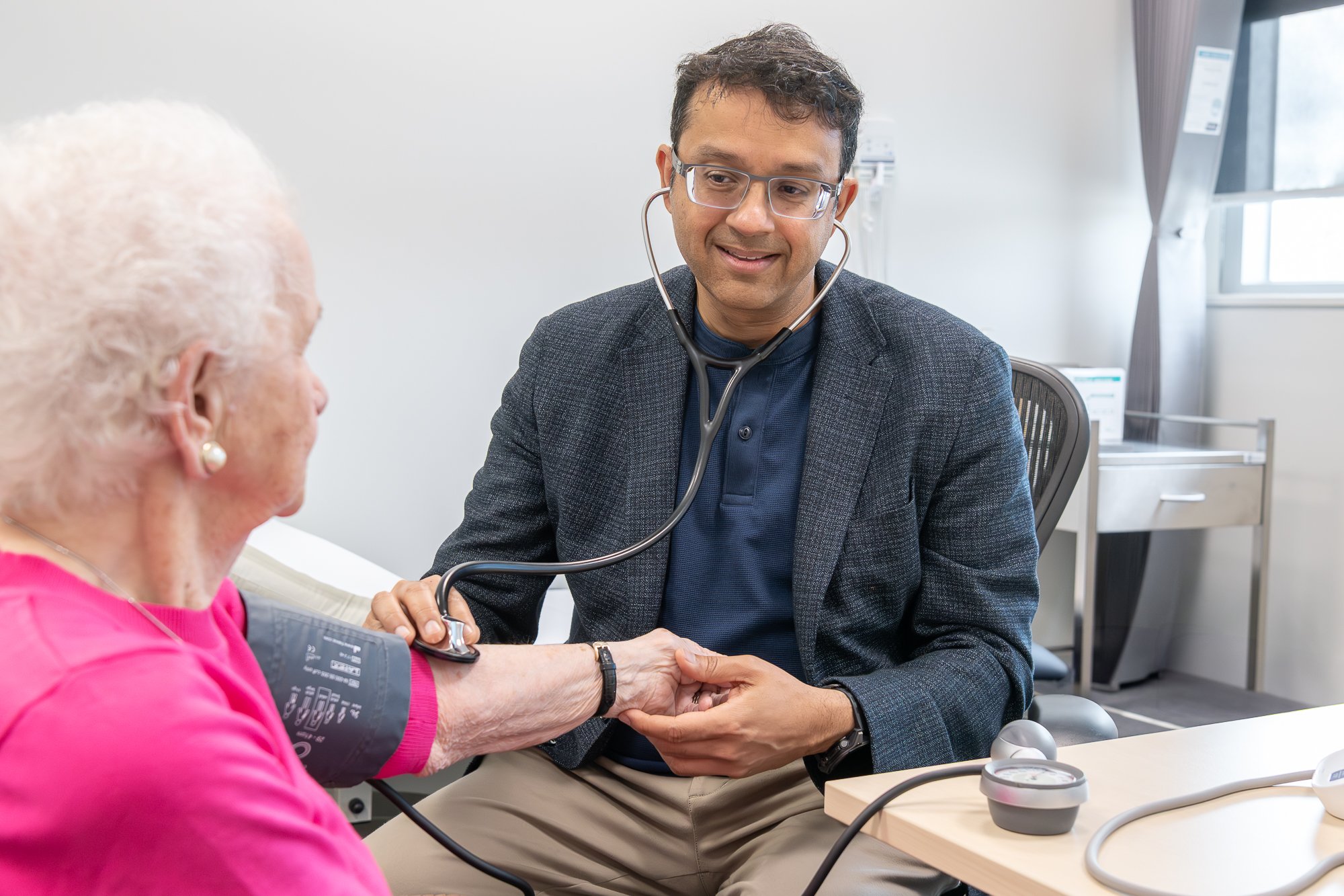 A young male doctor with glasses and a stethoscope around his neck examining an elderly woman in a bright, modern medical office.