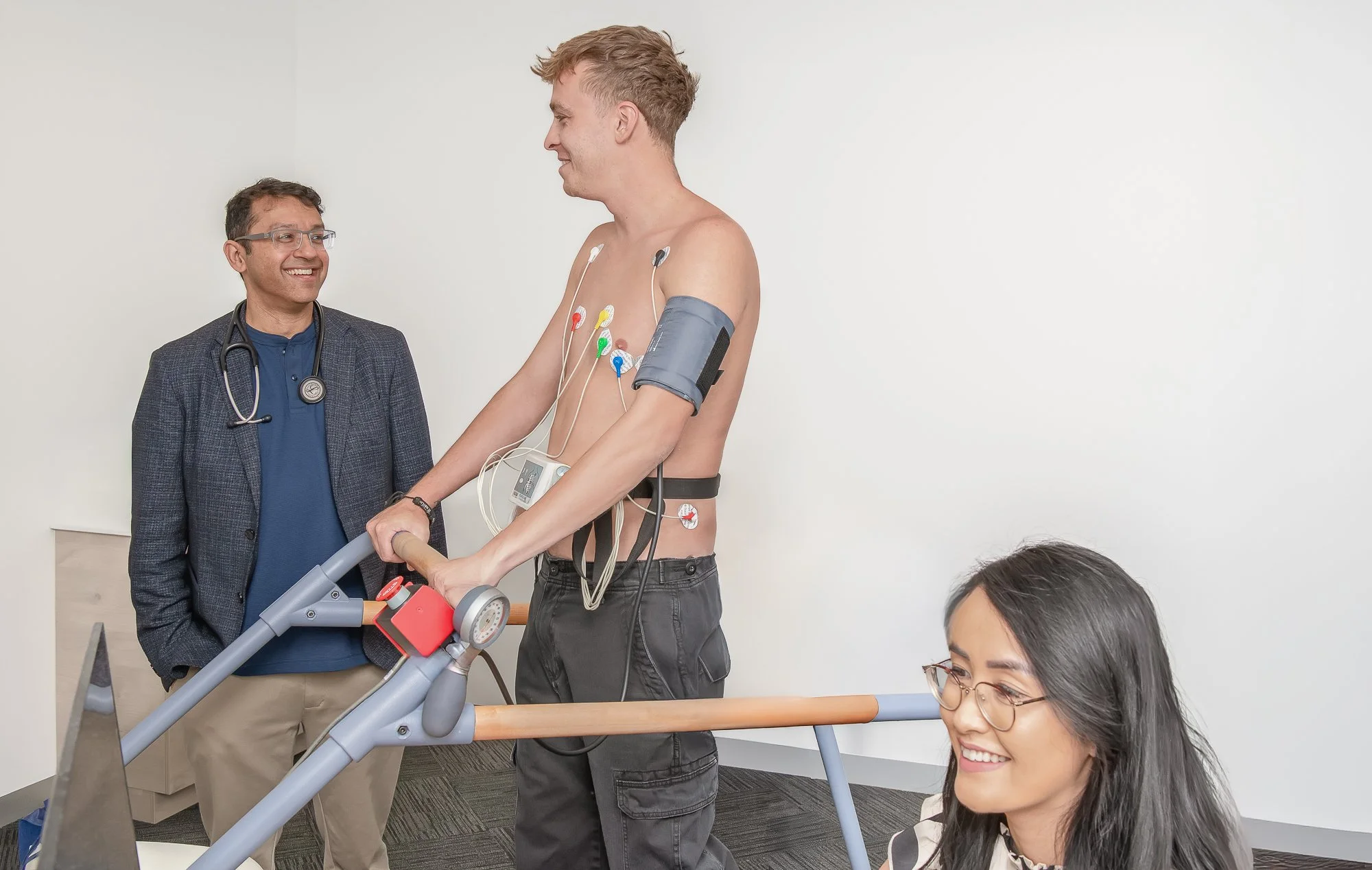 A male patient with electrodes attached to his chest is sitting on a treadmill, holding the handrails, in a medical examination room. A male doctor and a female patient are present, with the doctor smiling at the patient. The female patient is sitting and smiling, wearing glasses, and observing the scene. The room has plain white walls.