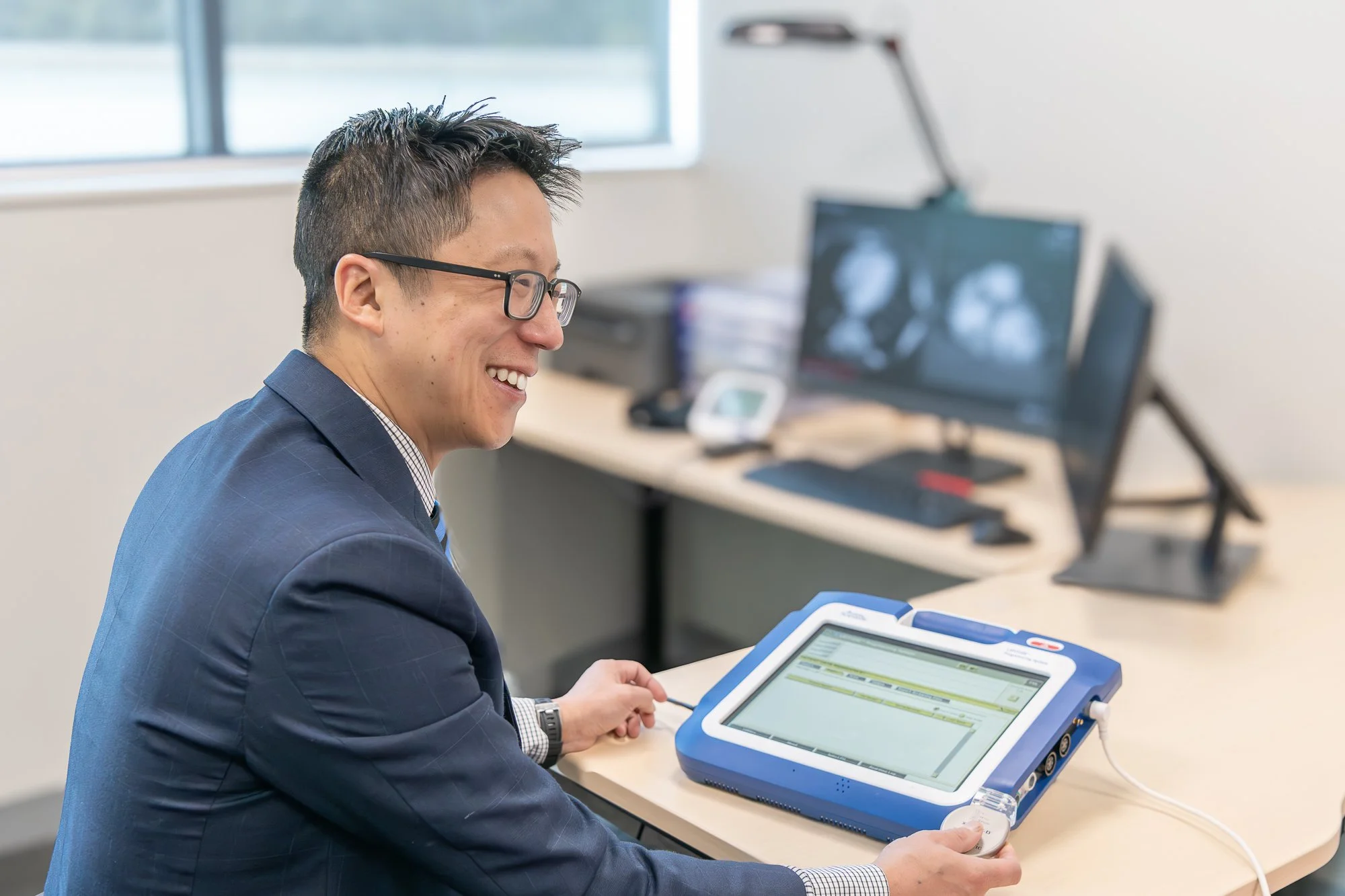 Smiling man in business attire sitting at desk using a medical or technical device with a screen in an office environment.