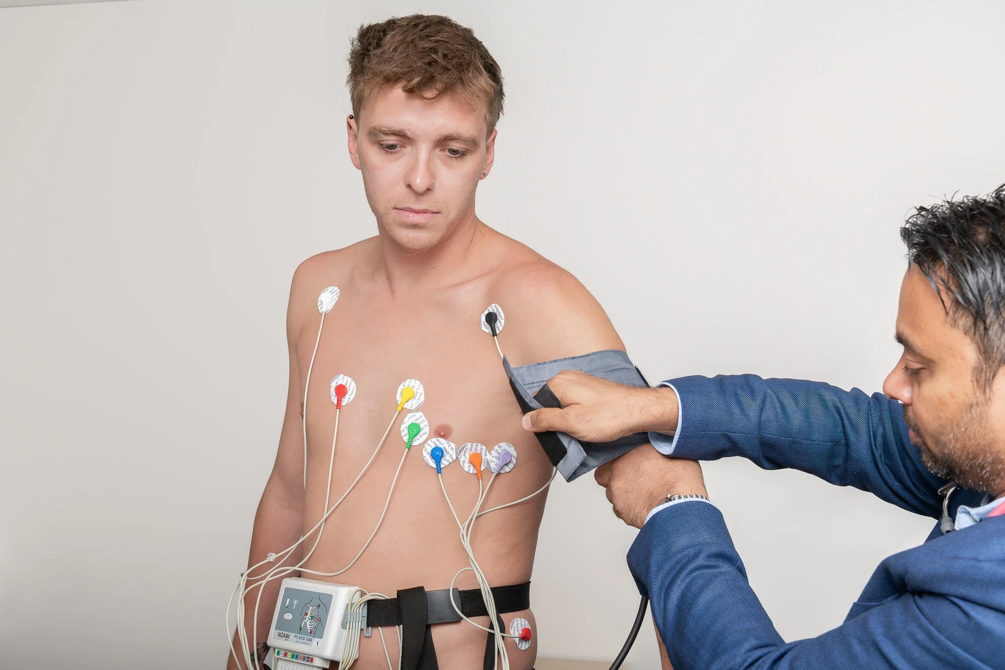 A young man undergoing an electrocardiogram test with multiple colorful electrodes attached to his chest, and a healthcare professional using a device to monitor his heart activity.