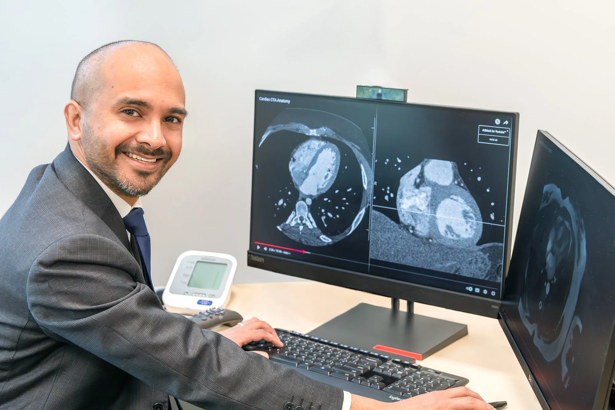 A man in a gray suit smiling while working at a computer with medical scans displayed on dual monitors, with a white medical device on the desk.