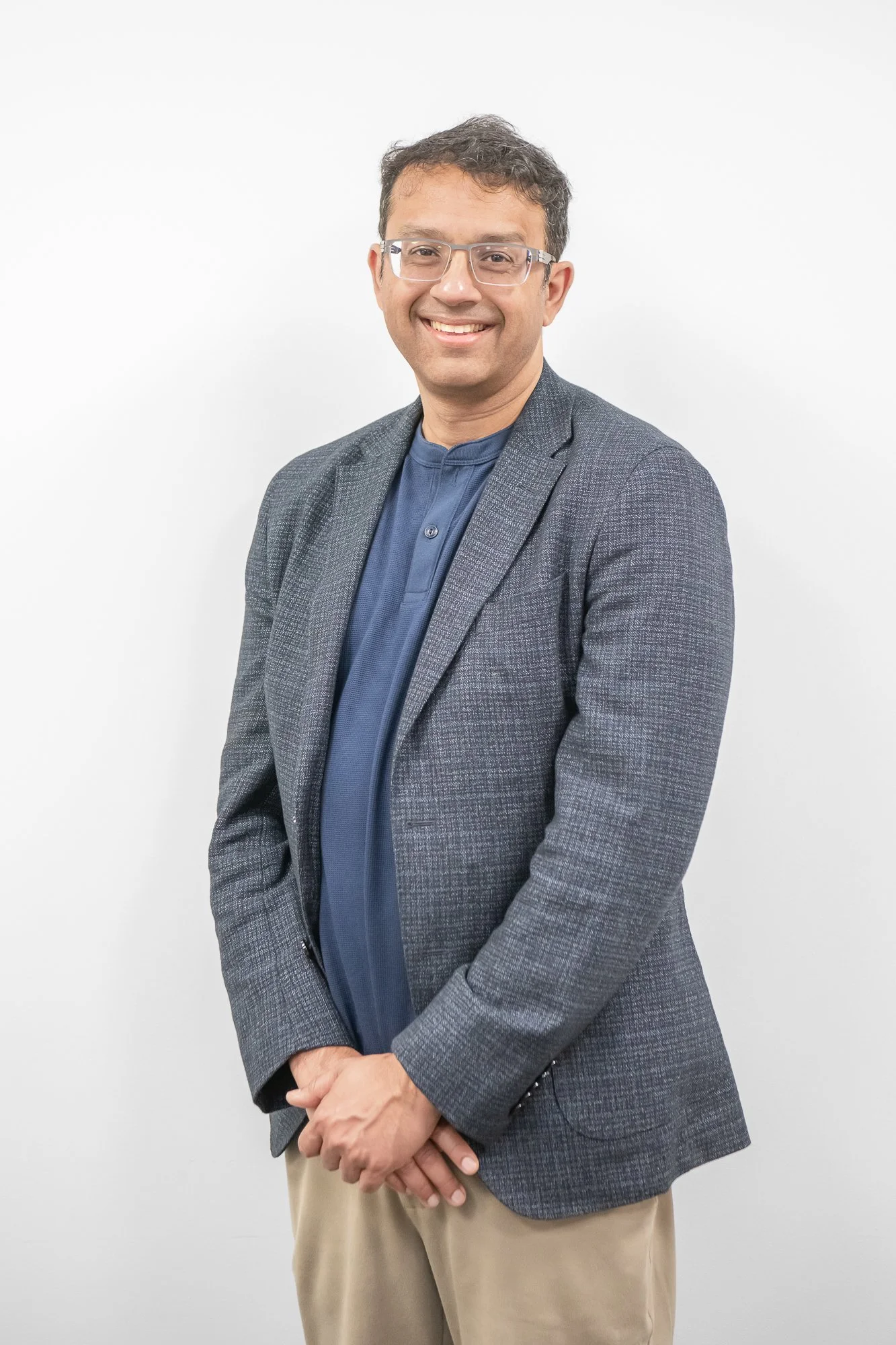 A man with glasses and a big smile is wearing a gray blazer, blue shirt, and beige pants, standing with hands clasped in front against a plain white background.