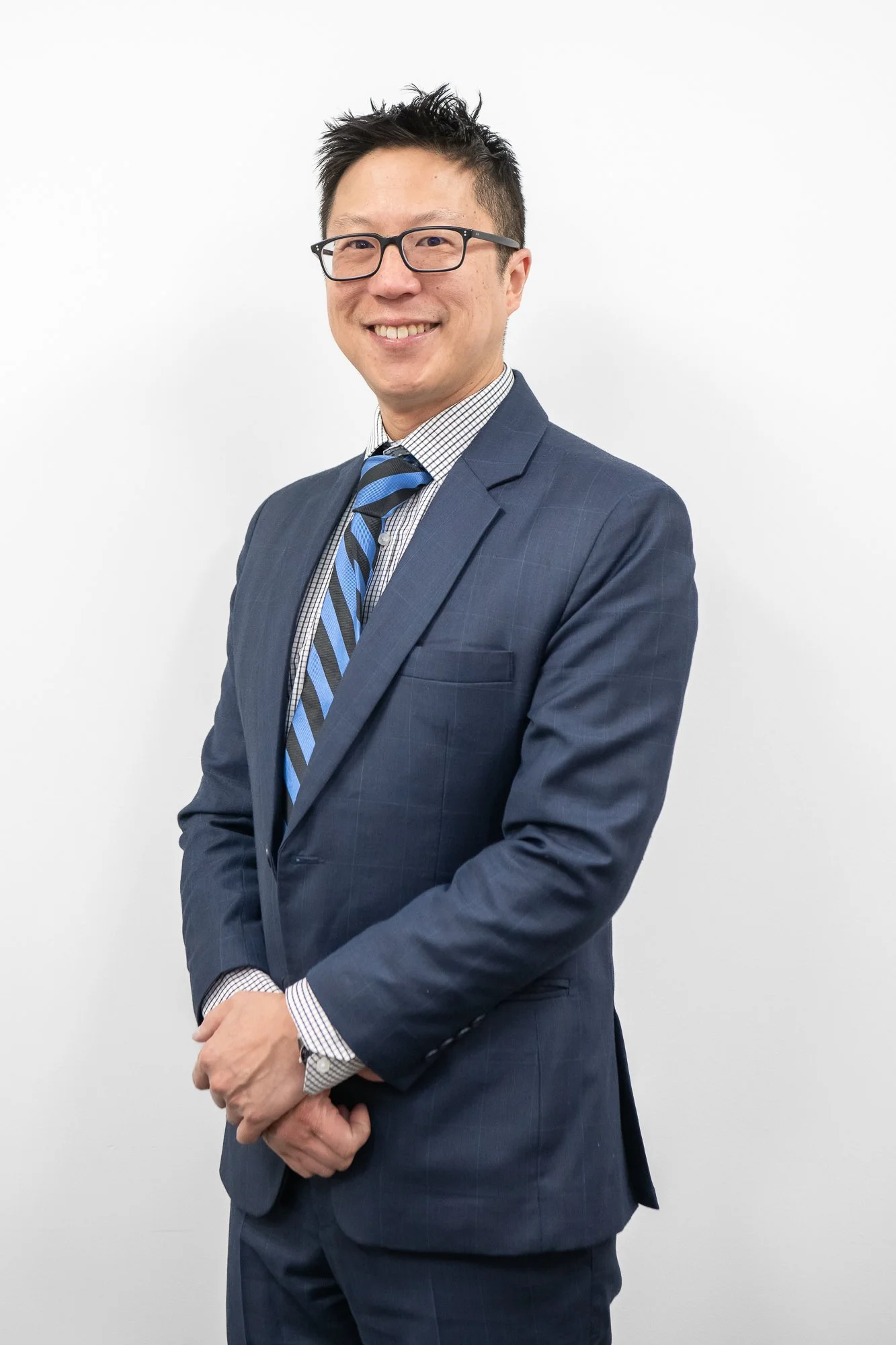 A professional man with black spiked hair, black glasses, and a friendly smile, dressed in a navy blue suit, light blue and white checkered shirt, and a striped blue and black tie, standing against a plain white background.
