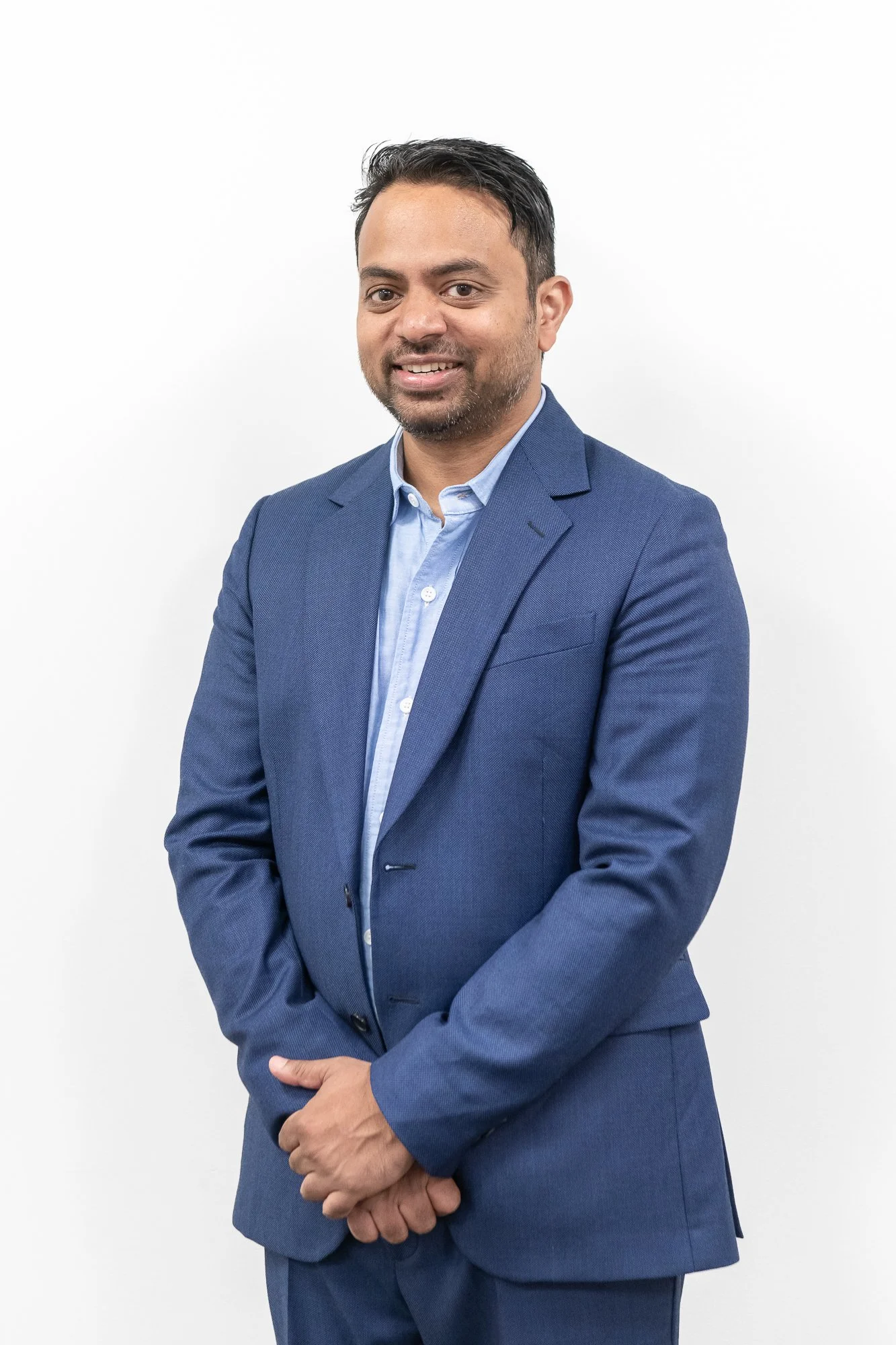 A man in a blue suit and light blue shirt standing against a plain white background.